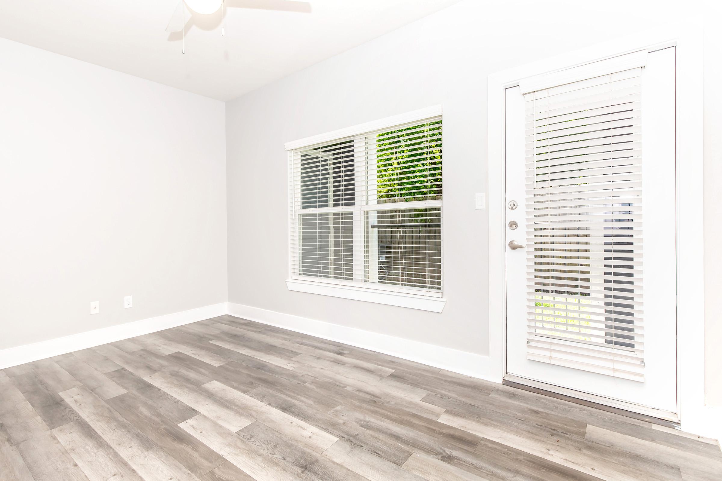 A bright, empty room with gray walls and light wooden flooring. There is a large window with white blinds allowing natural light in, and a door with slatted blinds leading outside. A ceiling fan is visible, enhancing the airy feel of the space.