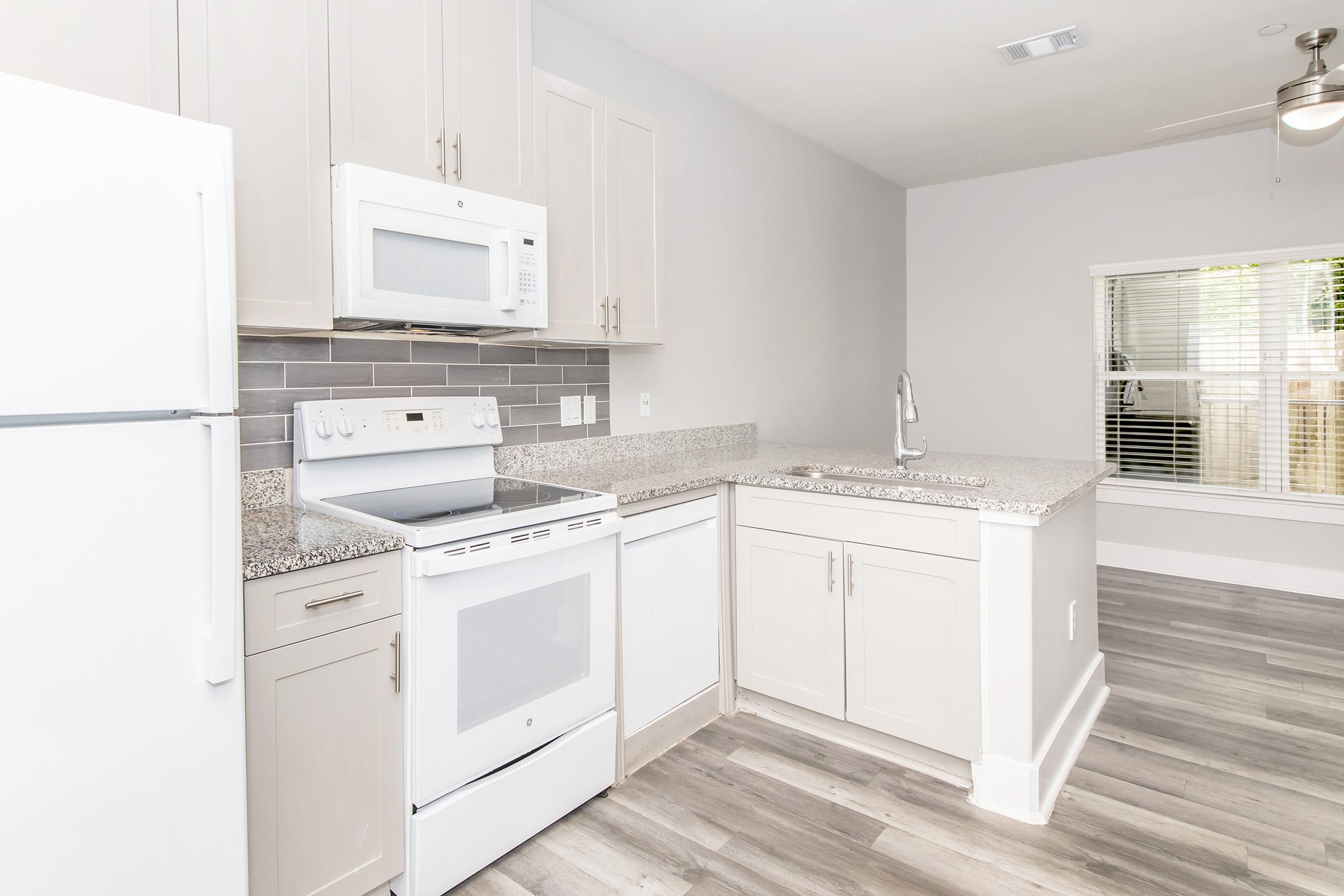 A modern kitchen featuring light-colored cabinets, a white refrigerator, a white oven with a microwave above, and a sink with a granite countertop. The space has gray tile backsplash and light wood-style flooring, with a window providing natural light.