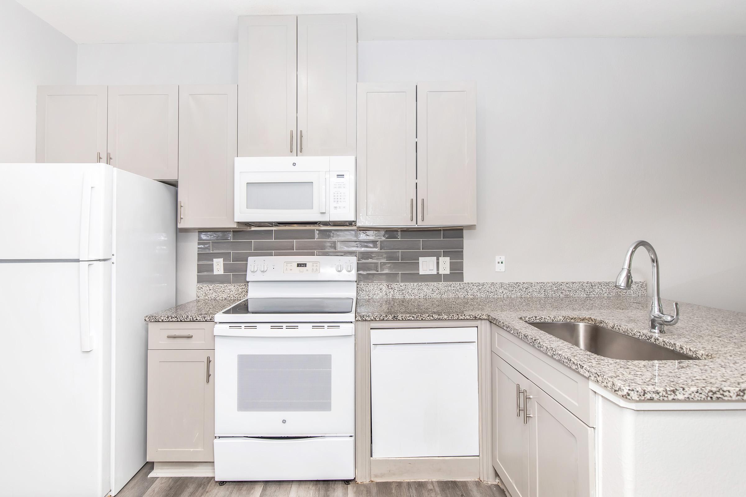A modern kitchen featuring light-colored cabinets, a white refrigerator, a stainless steel oven, and a microwave. The countertop is made of speckled granite, and there is a silver sink. The backsplash consists of gray tiles, creating a clean and stylish appearance.