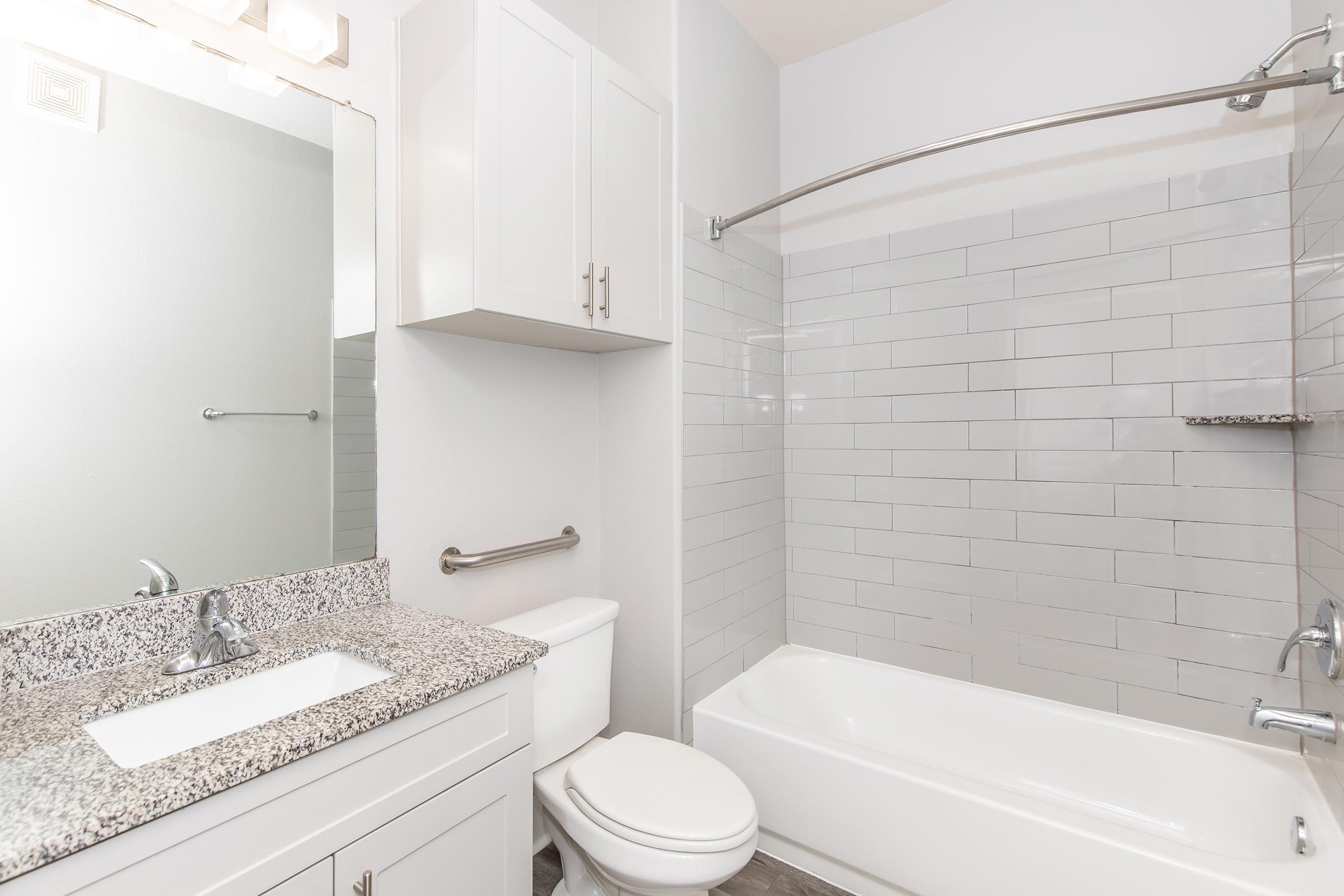 A modern bathroom featuring a white bathtub and shower, complemented by a granite countertop sink and a large mirror. The walls are tiled with light gray rectangles, and there is a white cabinet for storage. A towel bar and a small shelf are also visible, adding to the functionality of the space.