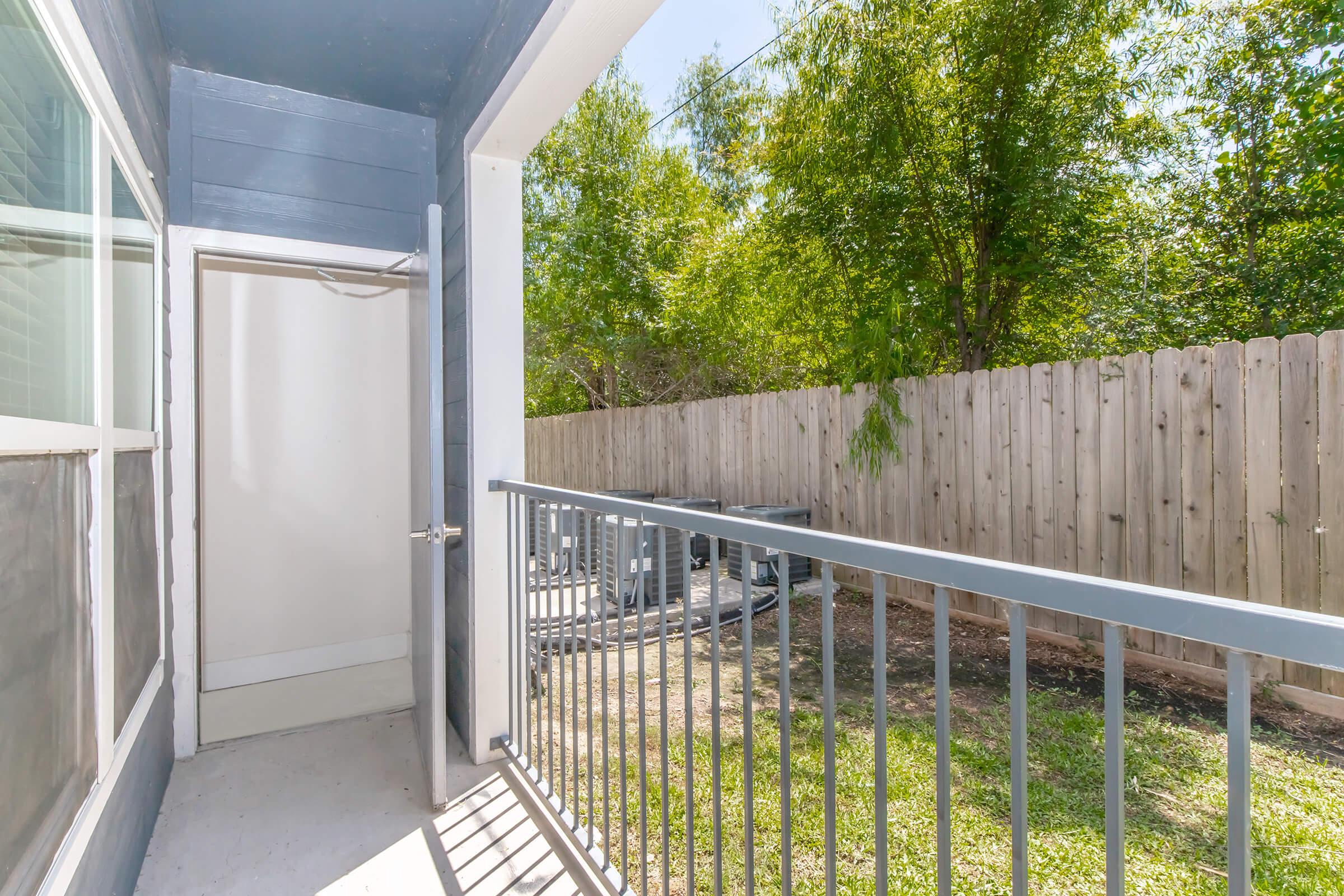 A view from a balcony overlooking a small outdoor area with a green lawn and fenced surroundings. The entrance to the interior is visible, along with a glimpse of outdoor equipment or furniture in the background. Natural light brightens the space.