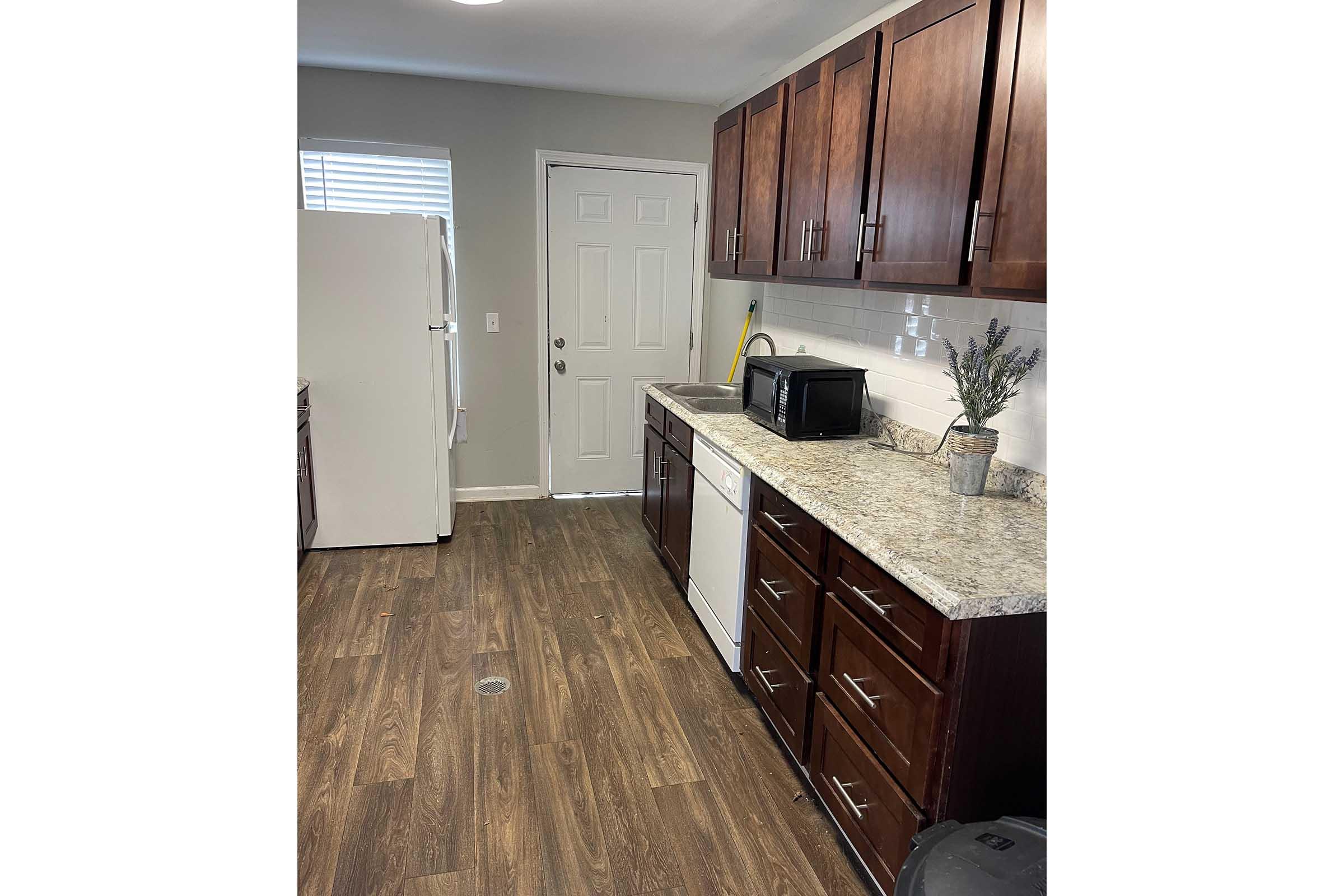 A modern kitchen featuring wooden cabinets, a countertop made of granite, and a white refrigerator. There is a silver microwave on the counter, alongside a small plant in a vase. The space has a light, neutral color scheme with window light streaming in. A door leads outside.