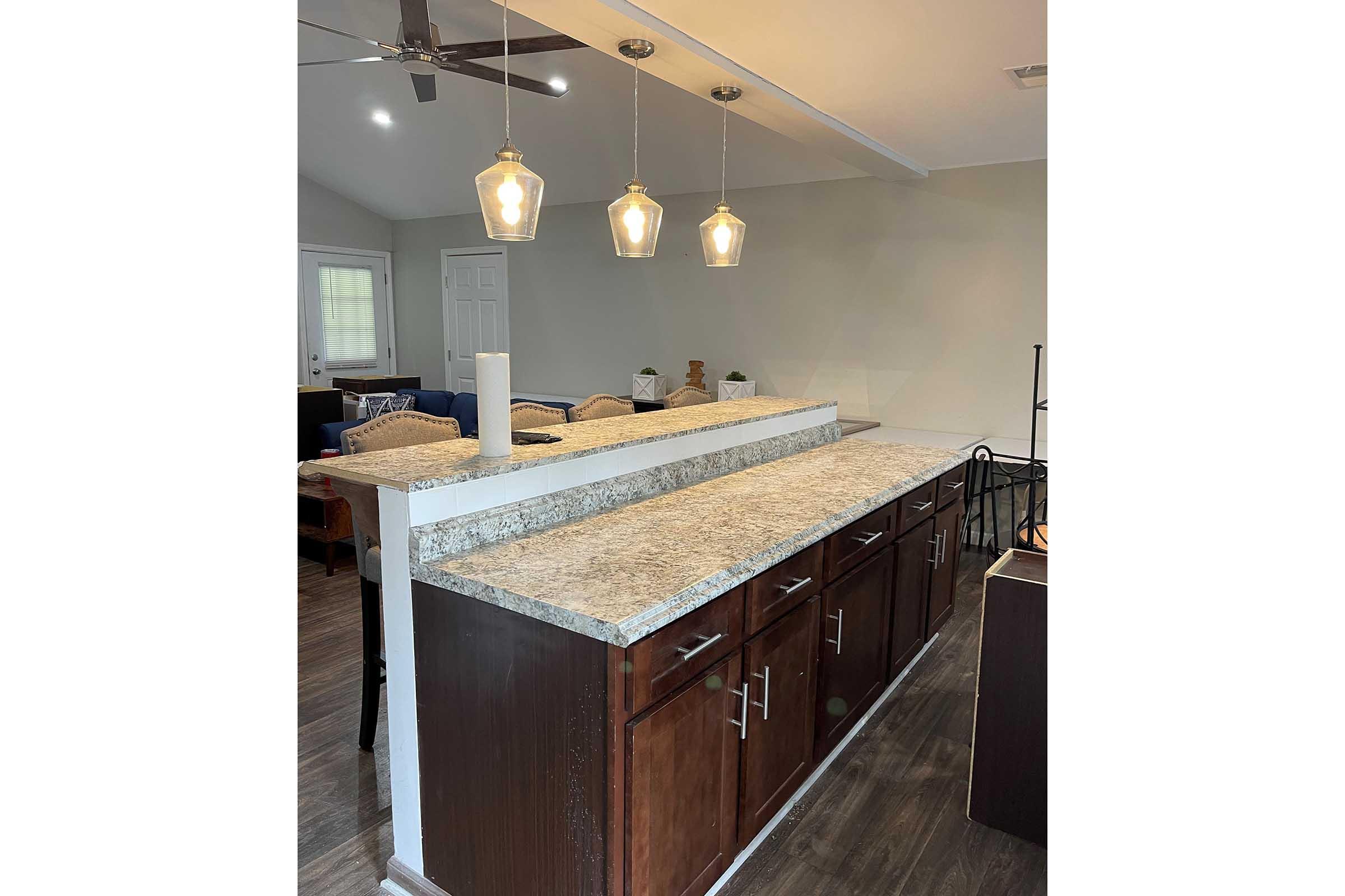 A kitchen with a long countertop featuring dark wood cabinets and a light-colored granite surface. Three pendant lights hang above the counter, and the space has an open layout with a seating area visible in the background. Natural light comes through a nearby window.