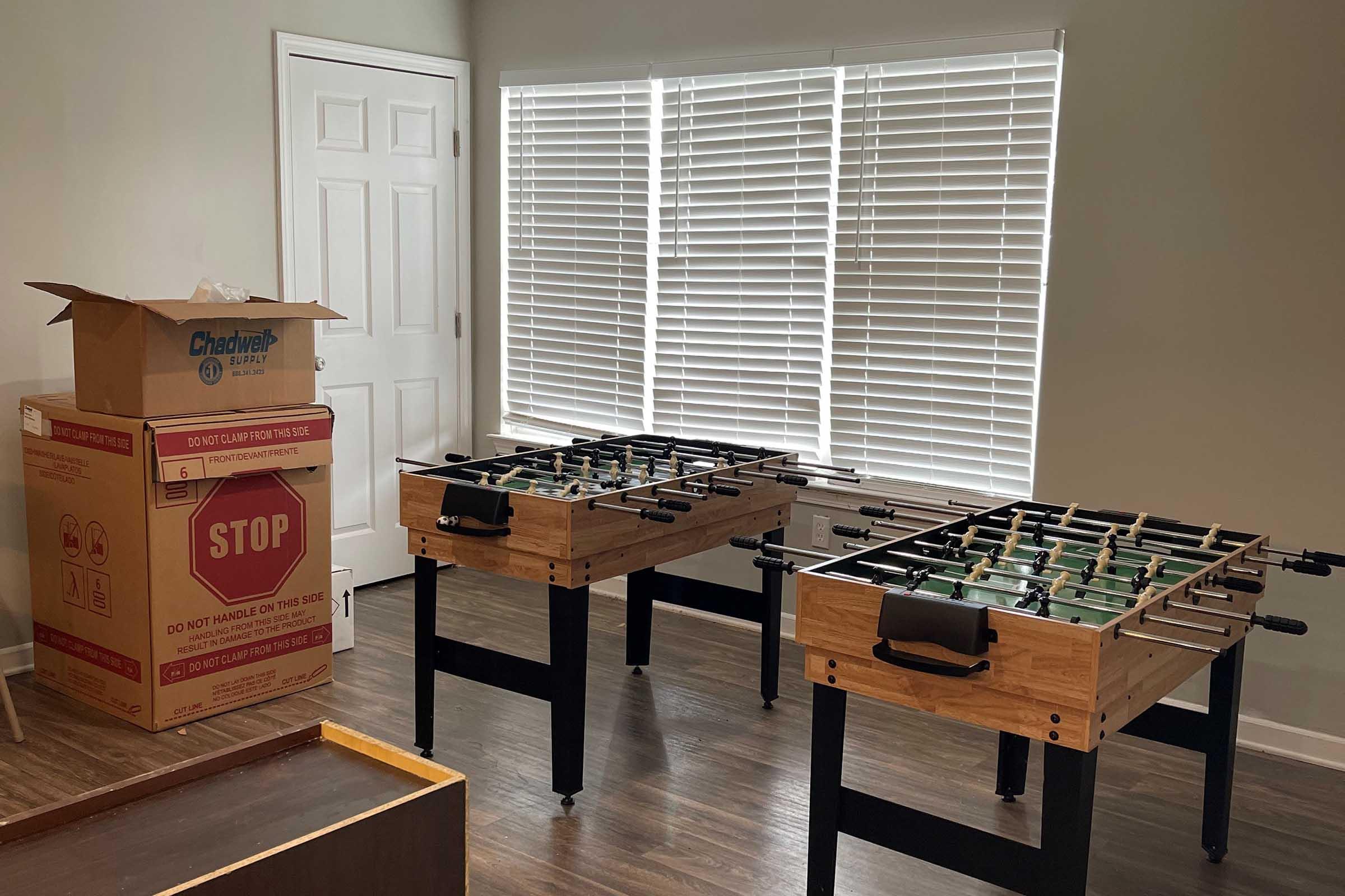 Two foosball tables are set up in a well-lit room with blinds, alongside a cardboard box marked "STOP" and a door partially visible in the background. The floor is wooden, and the overall atmosphere suggests a recreational space.