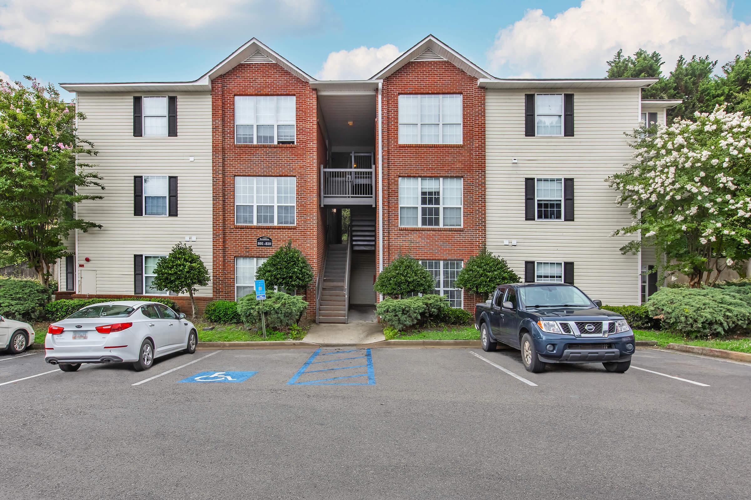 A multi-story apartment building with a combination of brick and siding exterior. The building has multiple windows and a central entrance. Surrounding the building are well-maintained shrubs and trees. Two cars are parked in front, including a white sedan and a dark pickup truck, with a designated handicapped parking space visible.