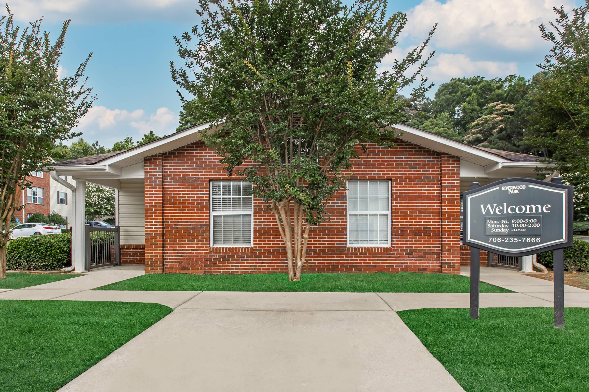 Brick building with a sign that reads "Welcome" outside. The entrance features a landscaped pathway with manicured grass and trees on either side. There's a contact number listed, and the scene includes clear blue skies and a lush background of trees.