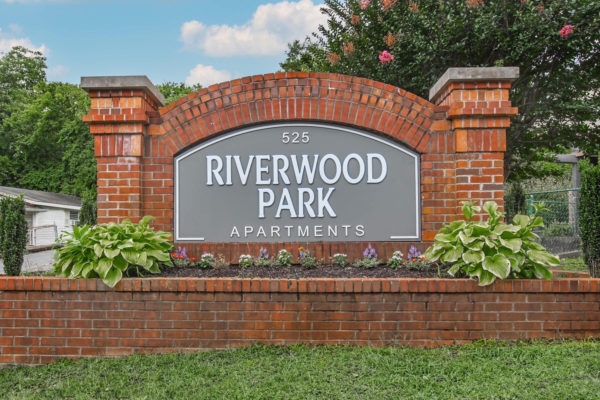 Sign for Riverwood Park Apartments at 525, featuring a brick structure with decorative plants at the base. The sign has a gray background with white text. The surrounding area includes green grass and trees in the background under a blue sky.