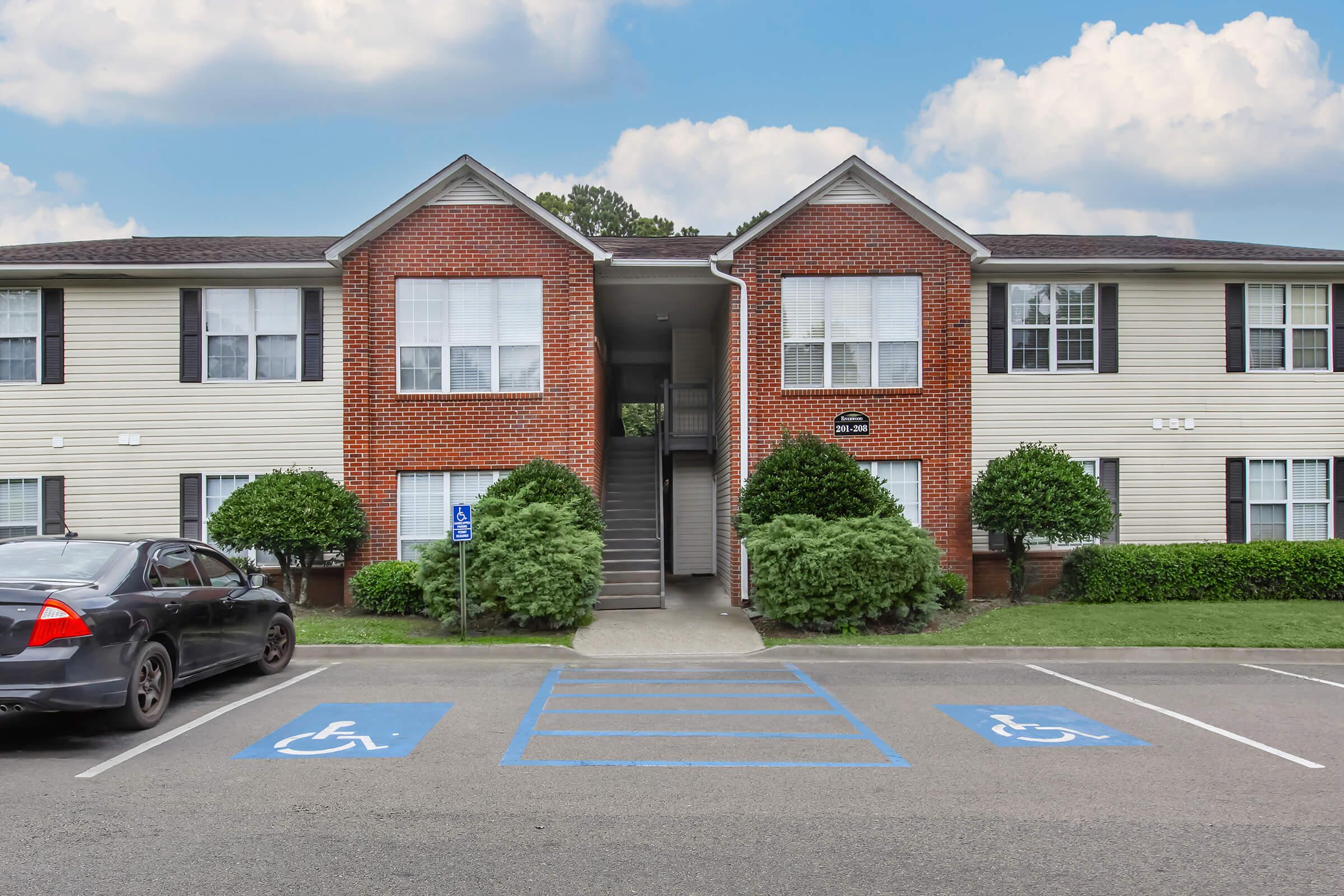 Front view of an apartment building featuring two stories with brick accents and large windows. There are accessible parking spaces in the foreground, marked with blue lines. Lush green shrubbery flanks the entrance on either side, contributing to the overall neat appearance of the property.