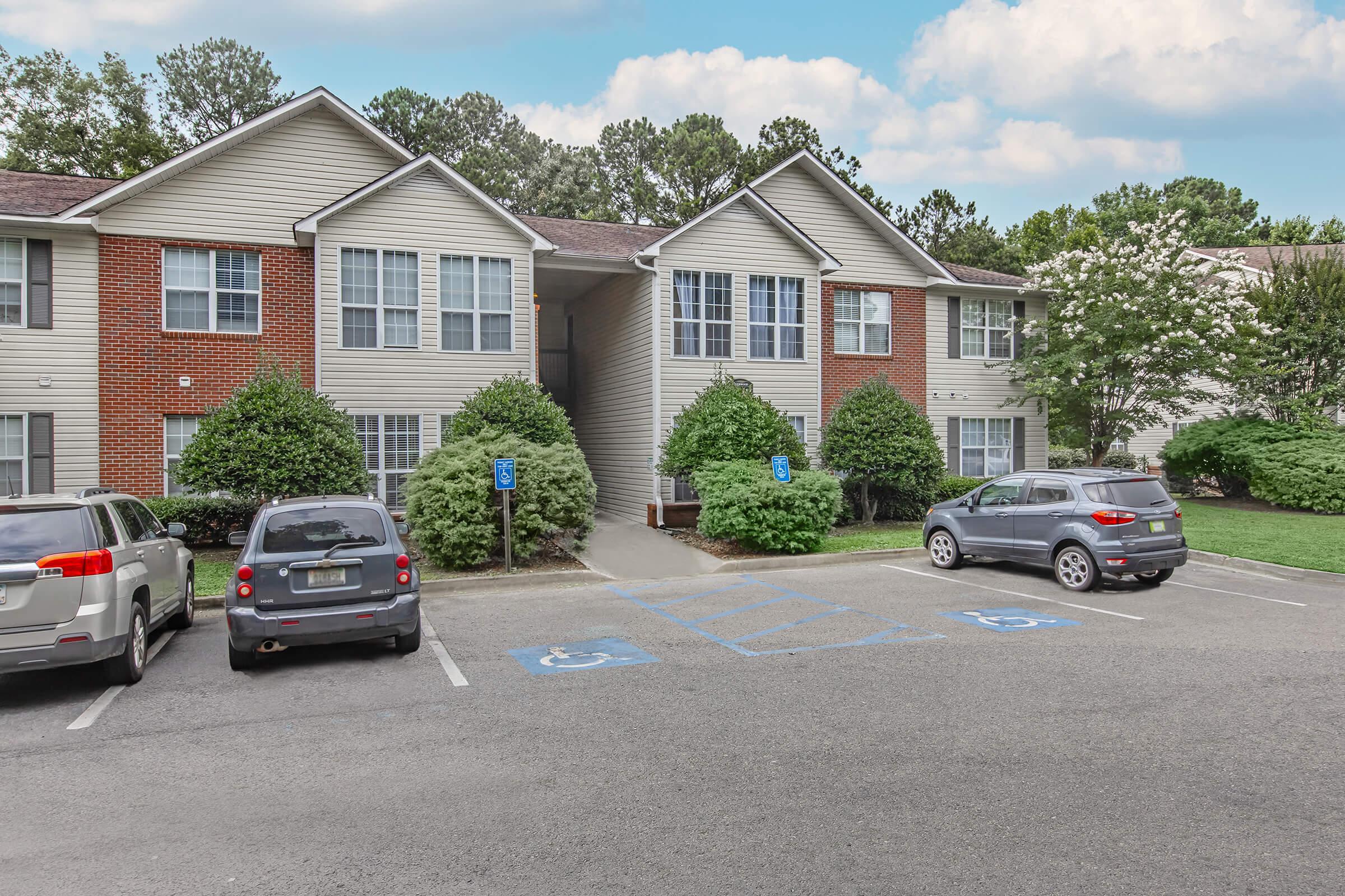 Two-story apartment building with a brick and siding exterior surrounded by green shrubs and trees. Parking area in the foreground with several parked cars, including two in designated handicapped spaces. The sky is partly cloudy, creating a bright atmosphere.