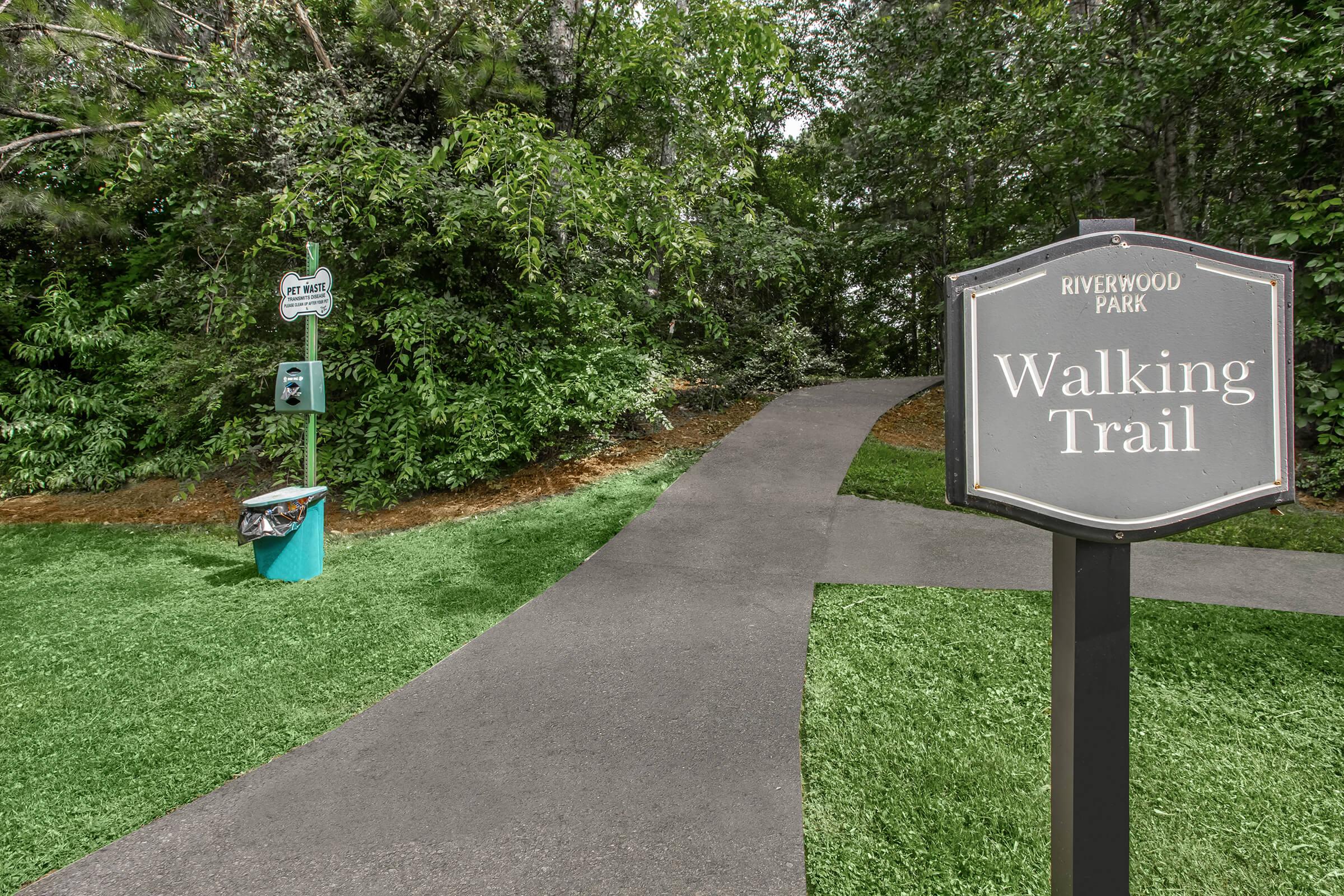 A signpost indicating a walking trail at Riverwood Park, surrounded by lush green foliage. The pathway is paved and well-maintained, leading into a natural, inviting area for walkers. There's also a nearby trash bin for convenience.
