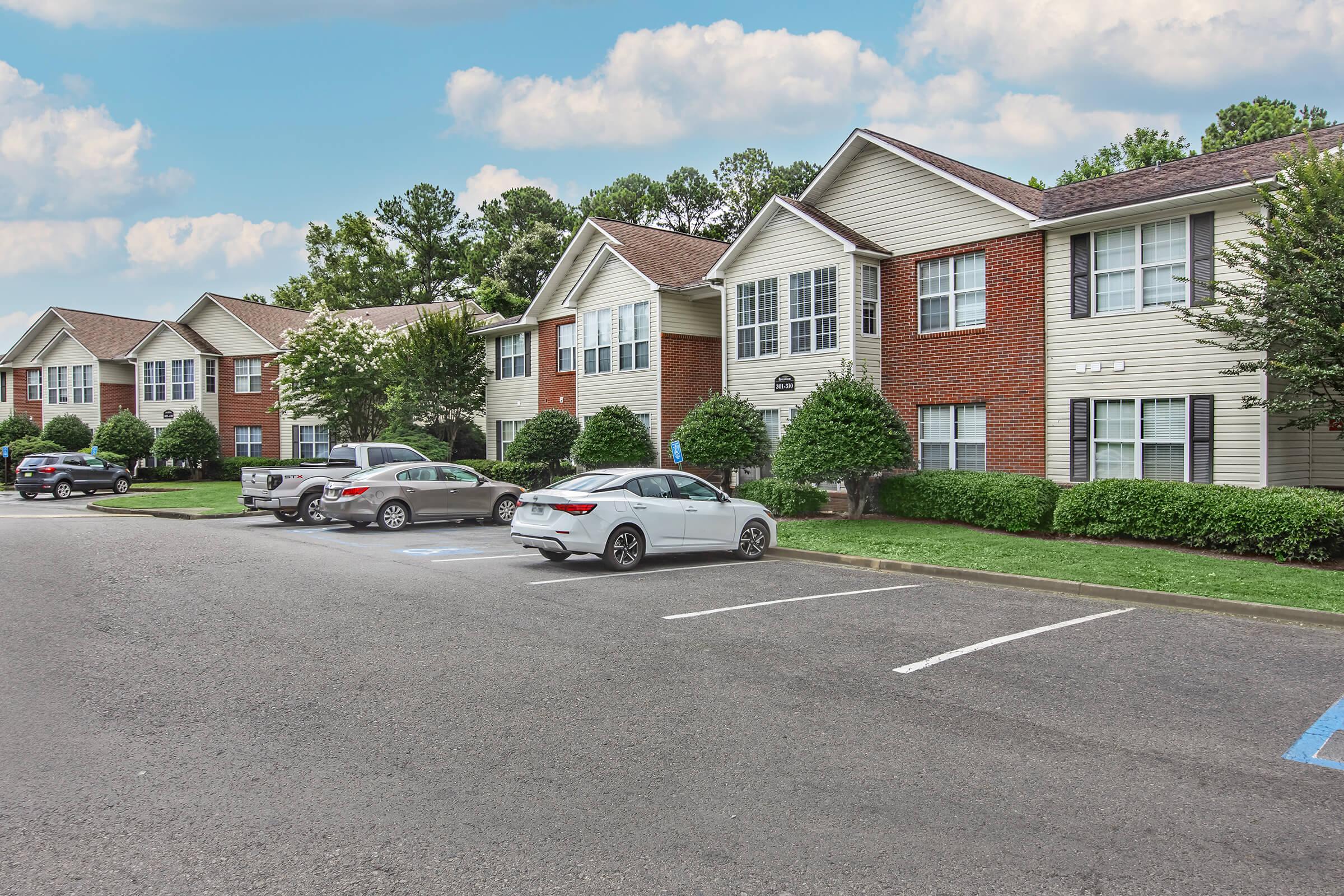 A view of a residential apartment complex featuring several two-story buildings, neatly landscaped lawns, and parked cars in the lot. The exterior includes a mix of brick and siding with green shrubs lining the pathway. The sky is clear with some clouds.