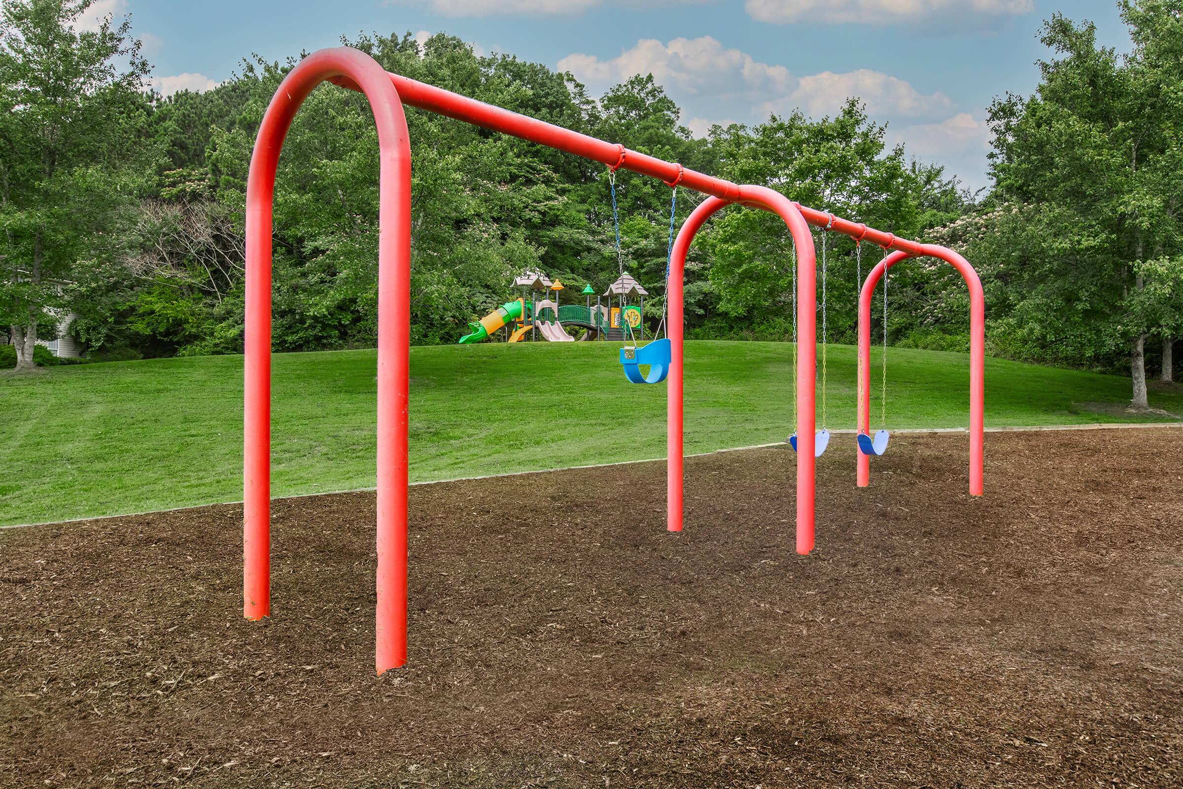 A playground scene featuring bright red swings with blue seats, set on brown mulch ground. In the background, there’s a green lawn with trees and a colorful play structure, including slides and climbing features, under a clear sky with fluffy clouds.