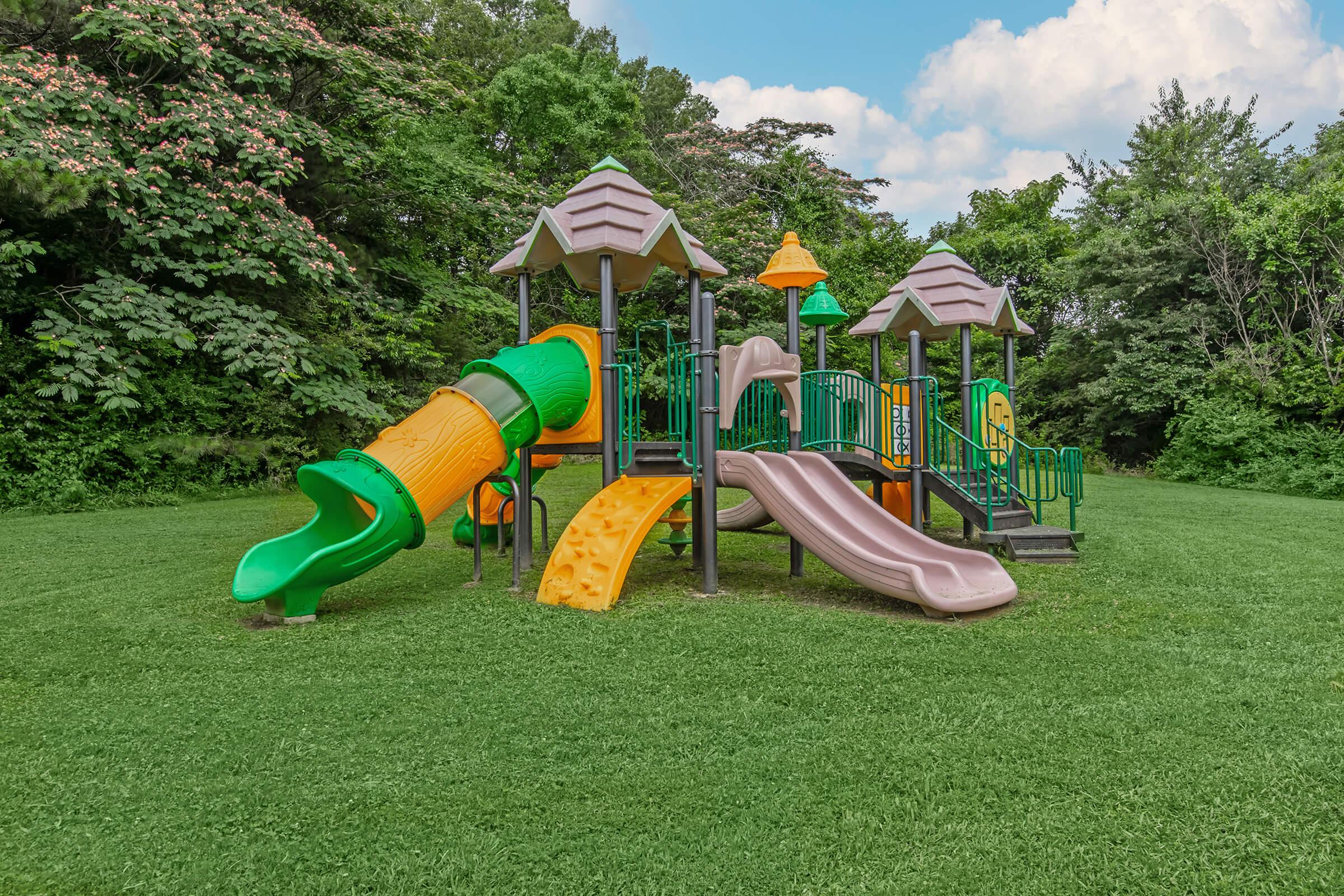 Colorful playground equipment featuring slides, climbing structures, and tunnels set on a grassy area surrounded by trees and blue sky. The design includes vibrant green, orange, and brown elements, inviting children to play and explore in a natural outdoor setting.