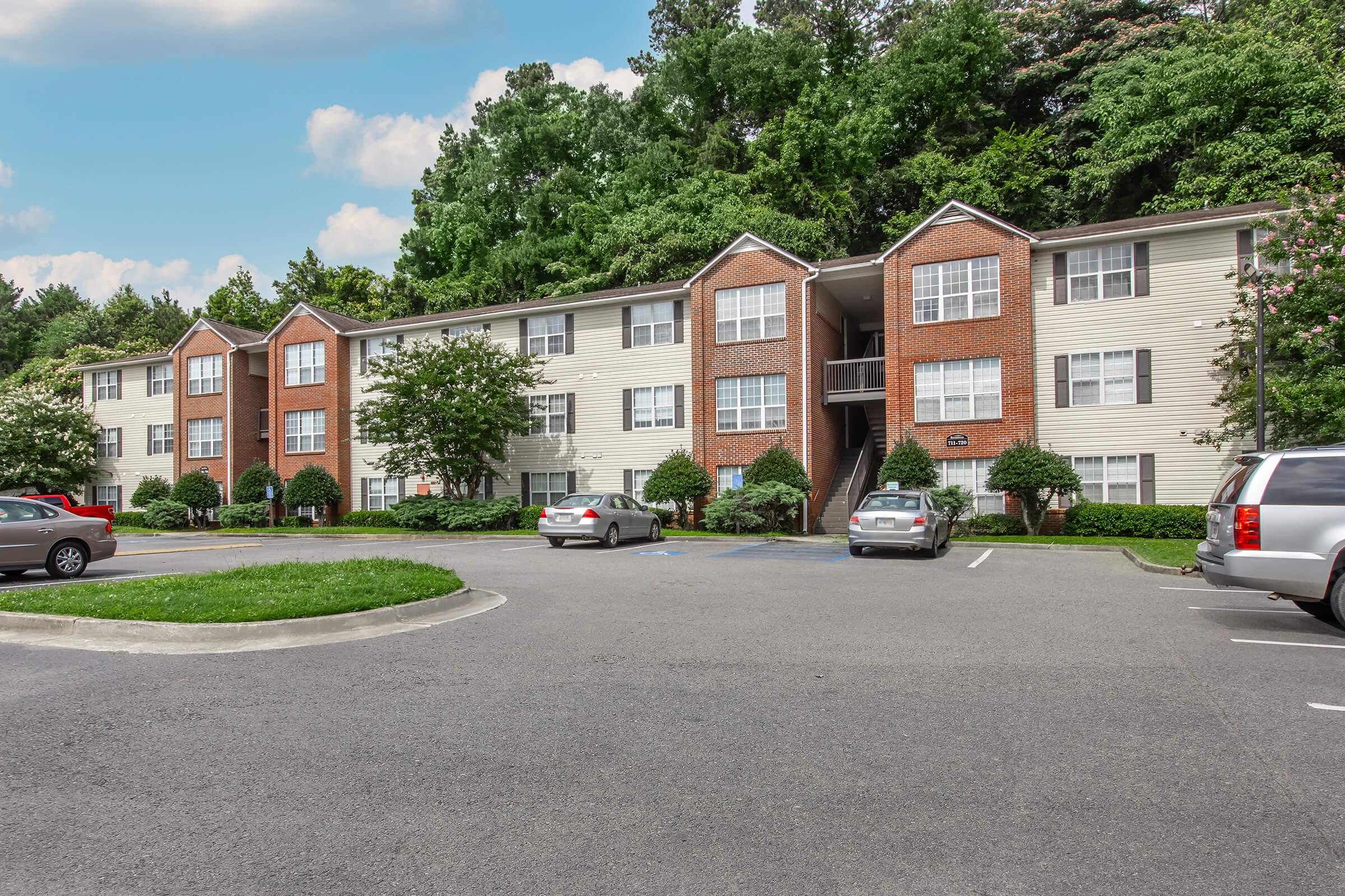 Three-story apartment building with a brick and beige facade, surrounded by lush green trees. A parking lot is in front, with several parked cars. The area is well-maintained, featuring landscaped shrubs and grass. The sky is partly cloudy, adding a bright ambiance to the setting.