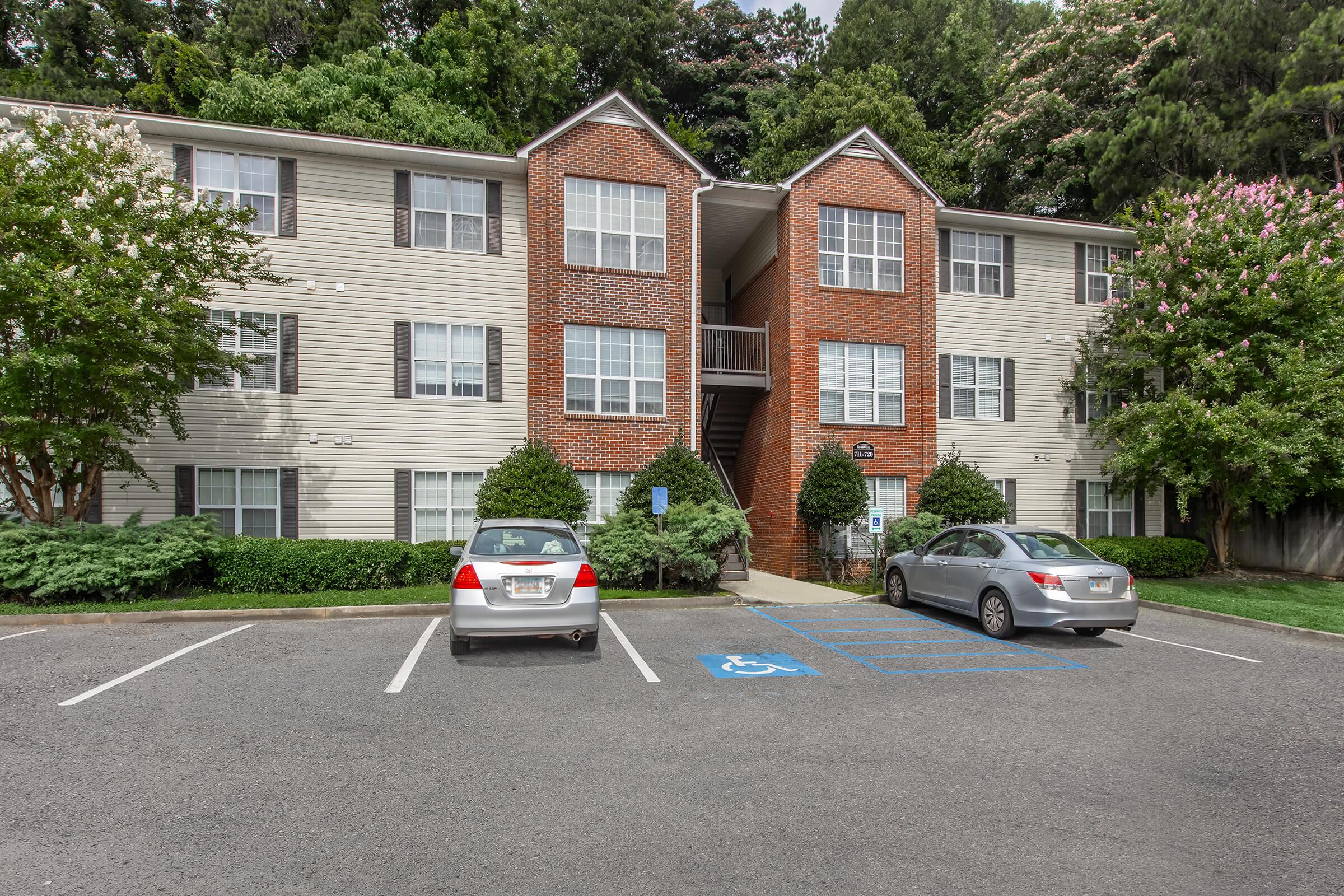 A three-story apartment building featuring a mix of brick and siding exterior. There are several windows on each floor, flanked by shrubs and trees. The parking area in front has two parked cars, one in a designated handicap spot, with a landscaped area in the background.