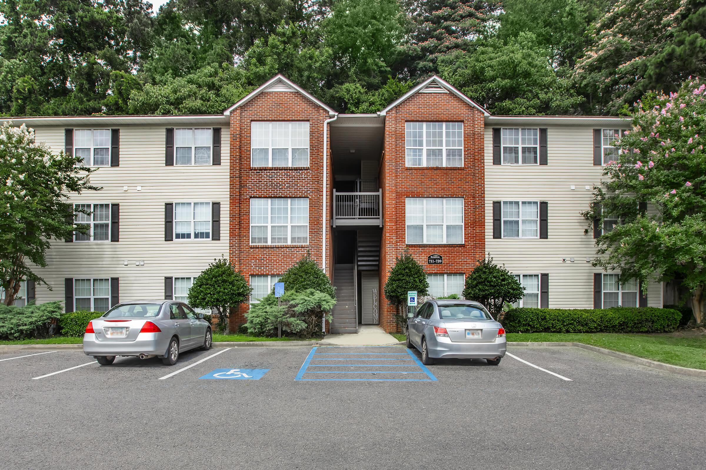 Three-story apartment building with a mix of brick and siding exteriors, surrounded by trees and shrubs. Two parked cars are visible in front of the building, one in a handicapped space. The building features multiple windows on each floor and a central entrance with stairs leading up to it.