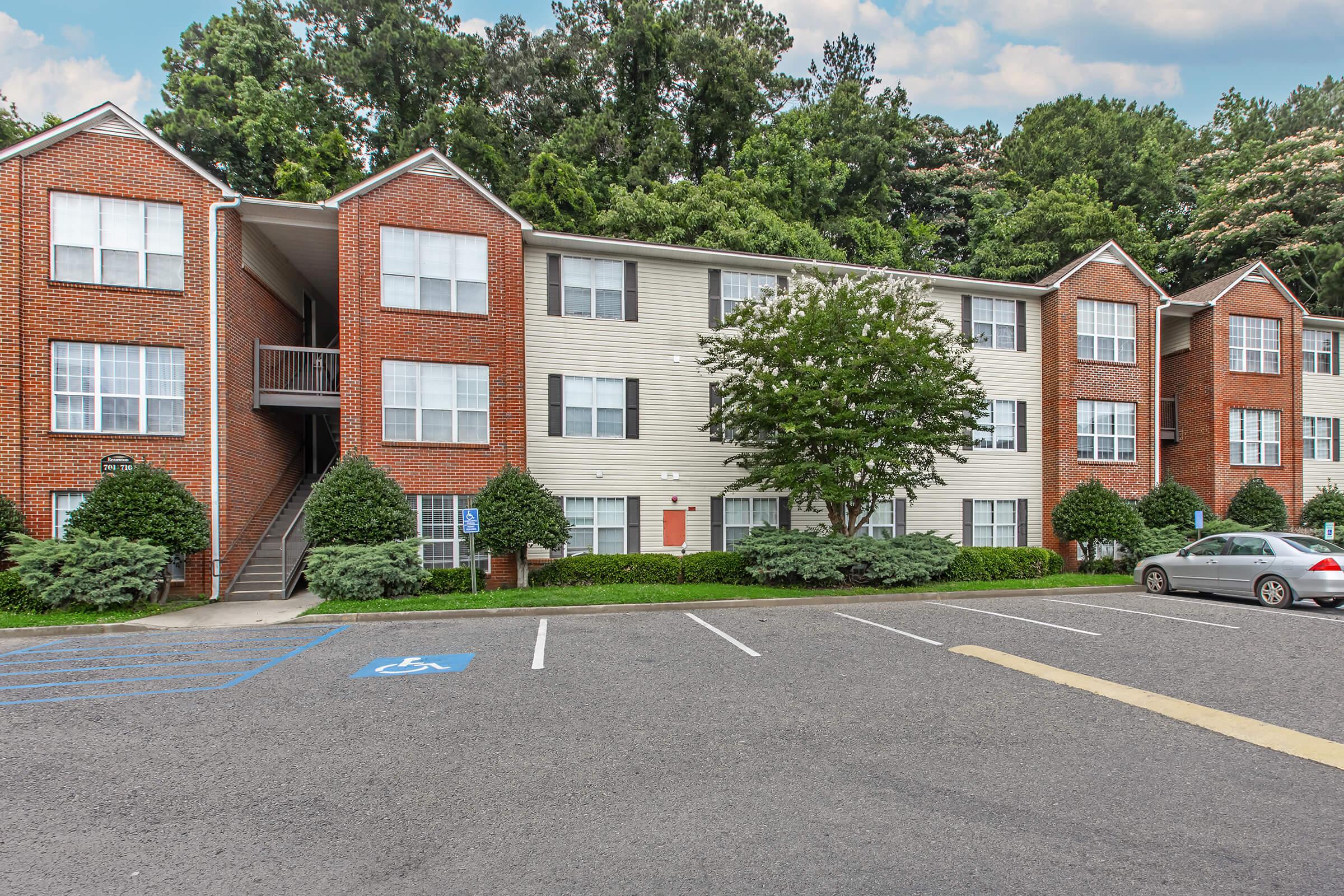 Three-story brick apartment building with white siding, surrounded by trees and shrubs. The building features multiple windows and a central entrance. A parking lot with designated handicapped spaces is in front, along with parked cars and a paved pathway leading to the entrance.