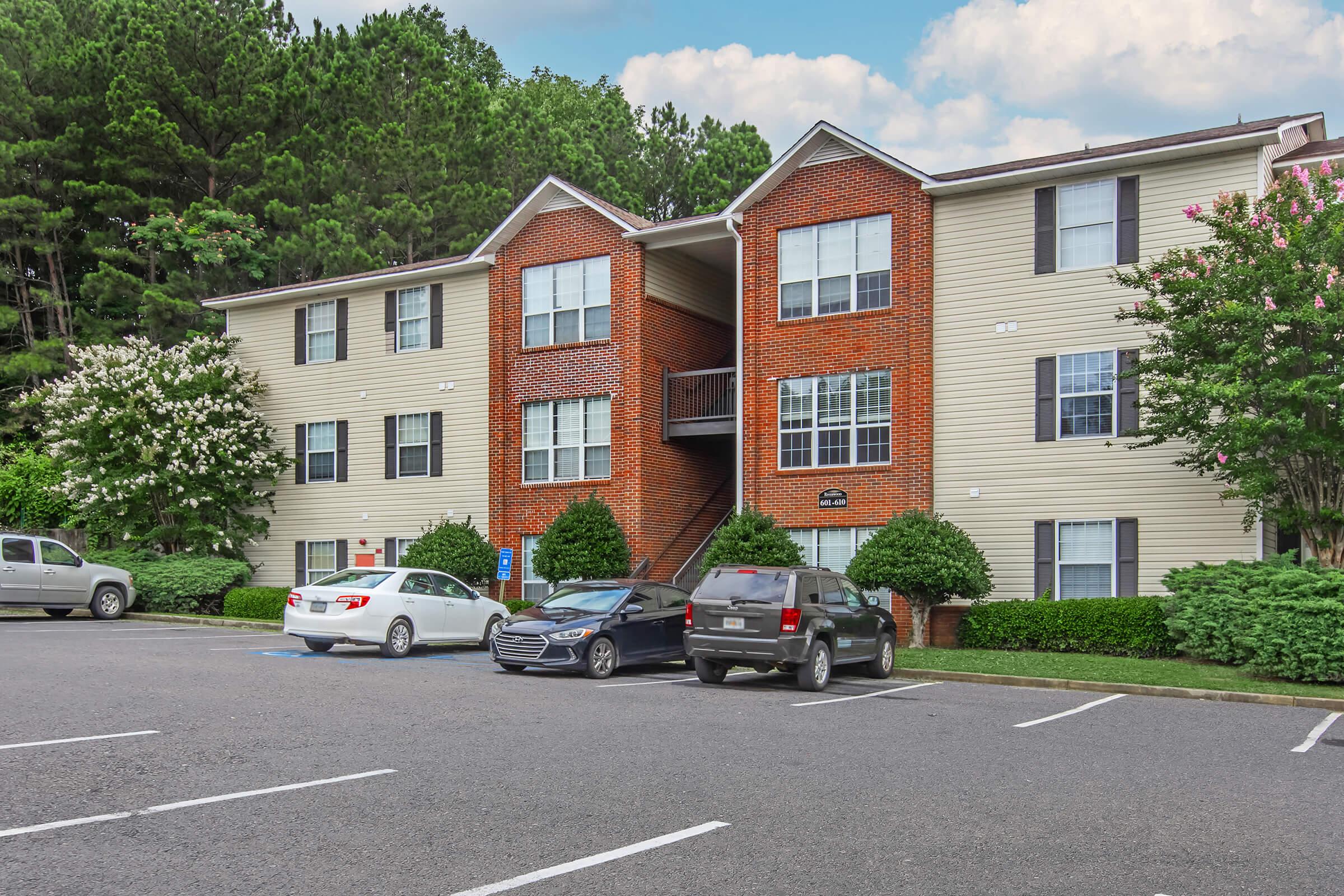 Three-story apartment building with a mix of brick and siding exterior. The front features multiple windows, a sign, and landscaped greenery. Several parked cars are visible in the foreground on a paved parking lot, with trees in the background under a partly cloudy sky.