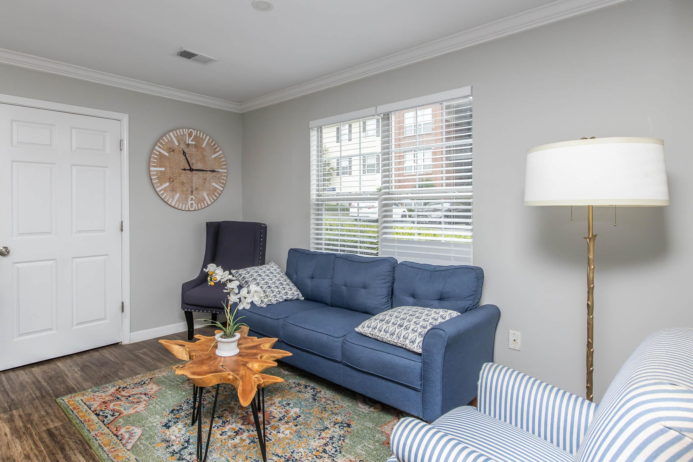 A cozy living room featuring a blue sofa with decorative pillows, a wooden coffee table, and a stylish lamp. There's a large clock on the wall and a plush armchair nearby. The room has light gray walls and a colorful area rug, with natural light streaming in through the window. A door leads to another space.
