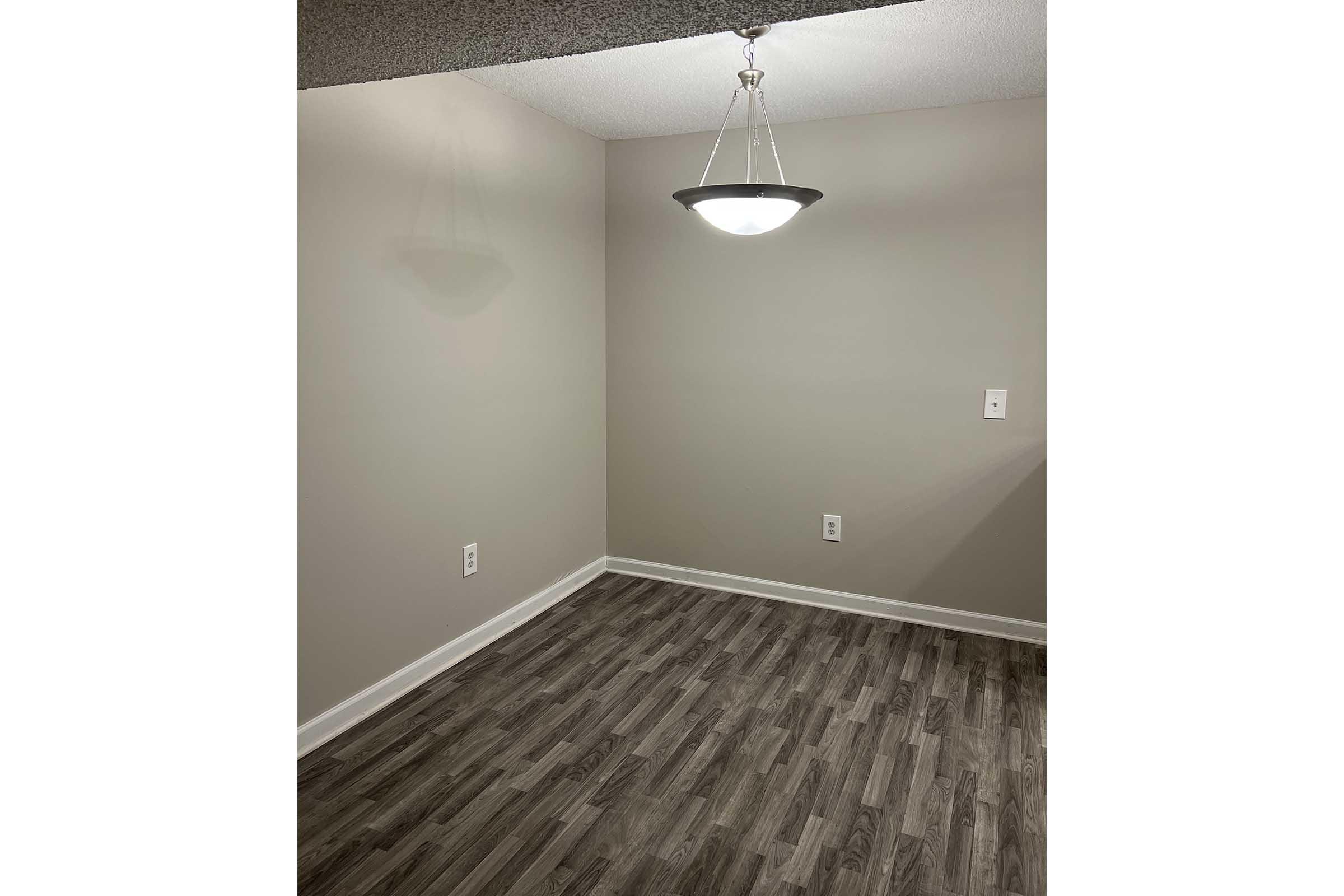 A small, empty room with light gray walls and a wooden floor. A round ceiling light hangs from the ceiling, illuminating the space. There are electrical outlets on the walls, but no furniture or decorations present. The overall ambiance is simple and minimalist.
