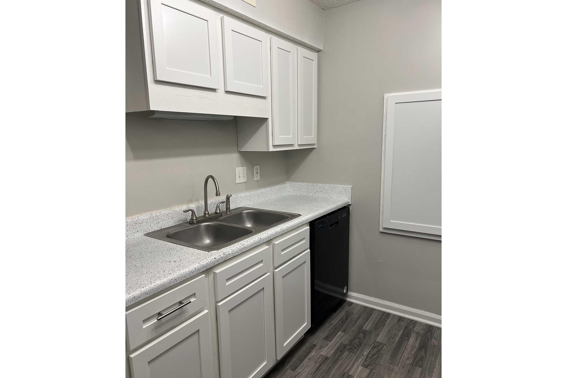 A modern kitchen featuring white cabinets, a double sink with a faucet, a black dishwasher, and a light-colored countertop. The walls are painted in a soft gray, and there is a space for a framed picture or bulletin board on the right side. The flooring is a wood-look laminate.