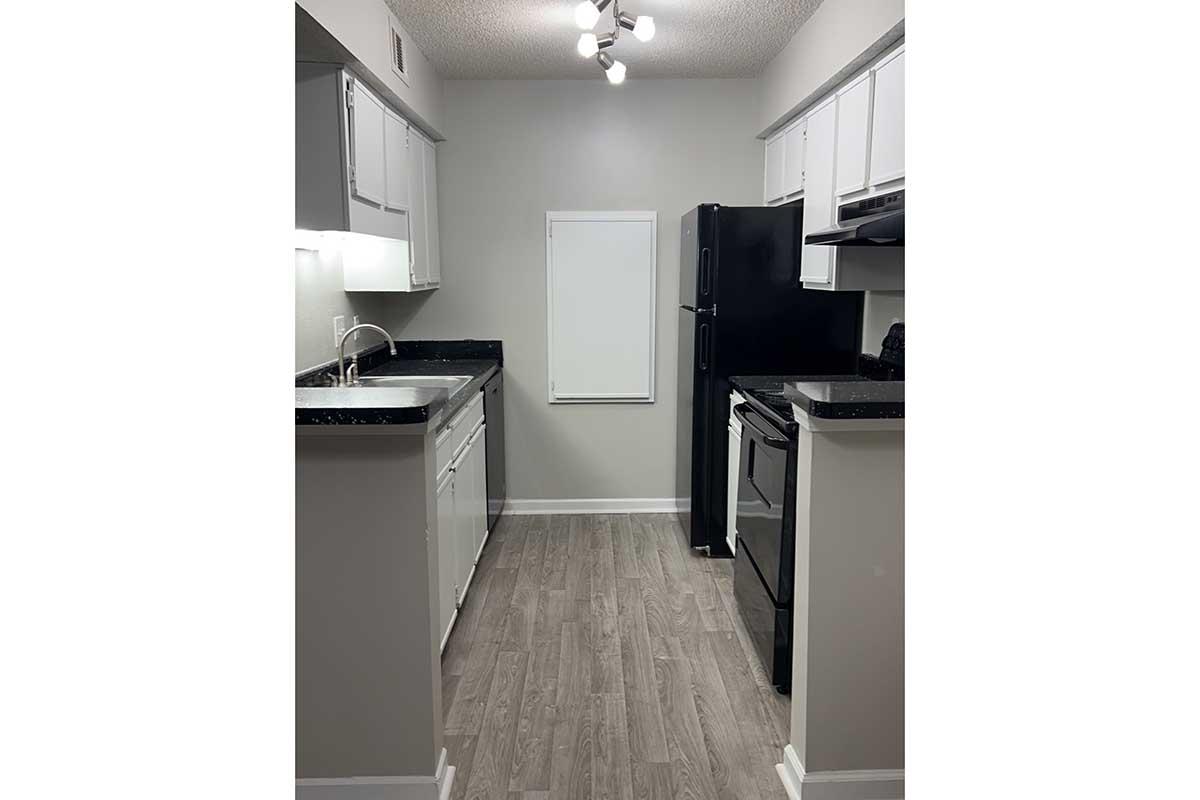 A modern kitchen featuring gray walls and wood-like flooring. The space includes white cabinets, a black refrigerator, a black stove, and a black sink with a chrome faucet. A window is partially covered, allowing some natural light into the area. The overall design is clean and minimalist.