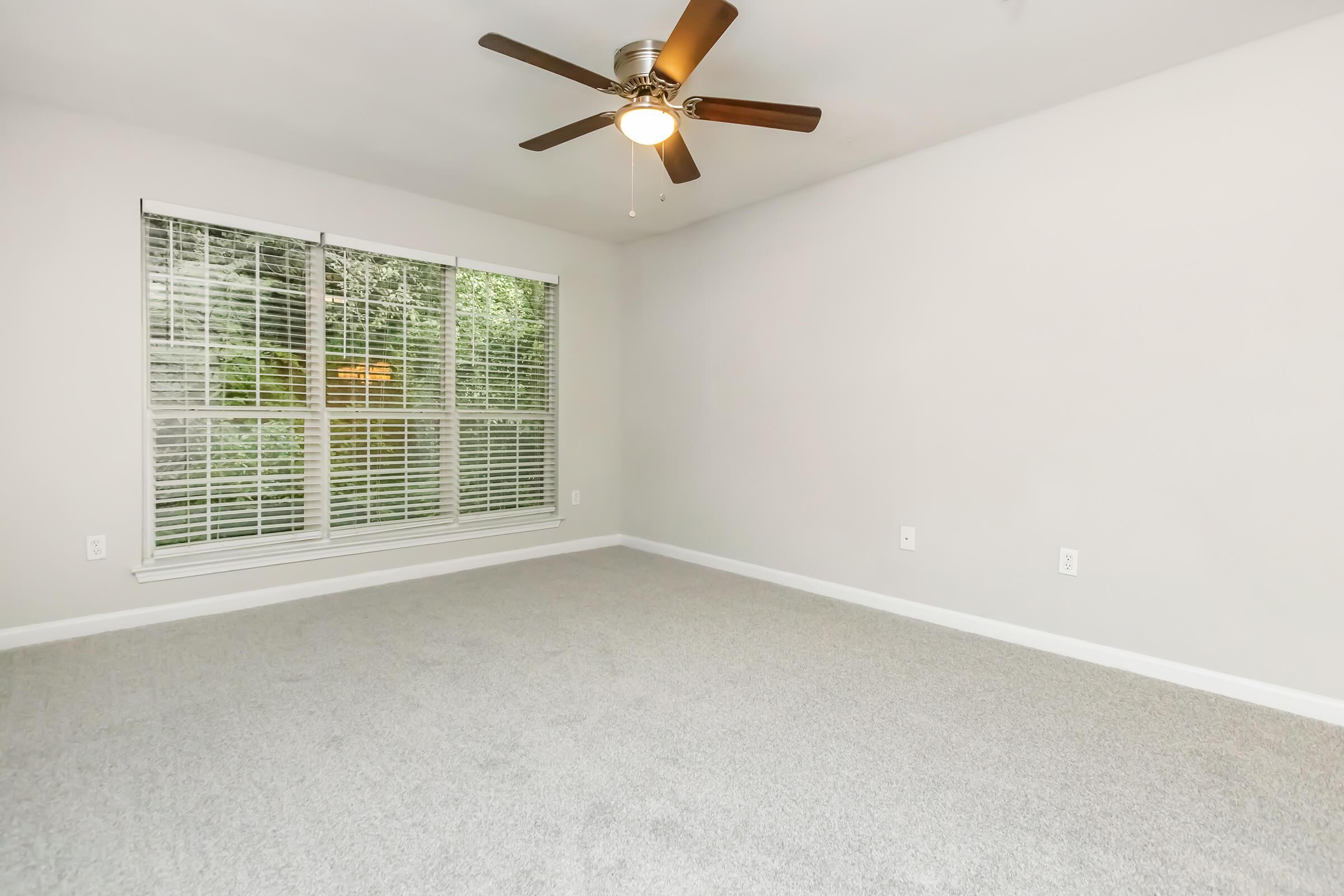 A spacious, empty room featuring light gray carpet, a ceiling fan with wooden blades, and large windows with white blinds that let in natural light. The walls are painted a light neutral color, creating a bright and airy atmosphere.