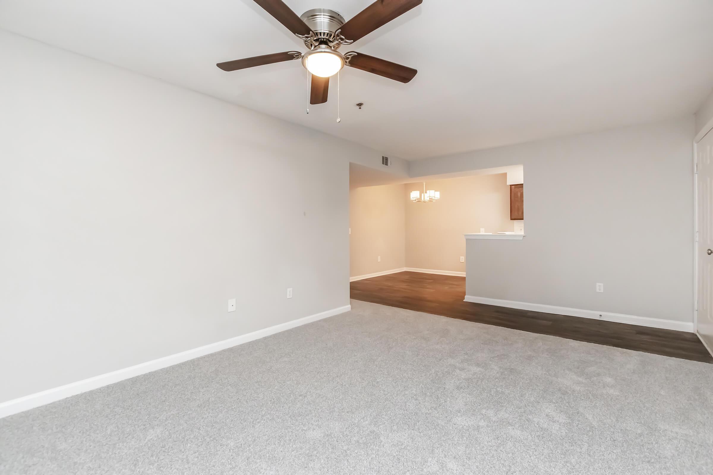 A spacious, empty living room featuring light gray walls, plush carpet, a ceiling fan with light, and an open layout leading to a kitchen area in the background with wooden cabinets and pendant lighting. The room is well-lit, offering a versatile space for furniture arrangement.