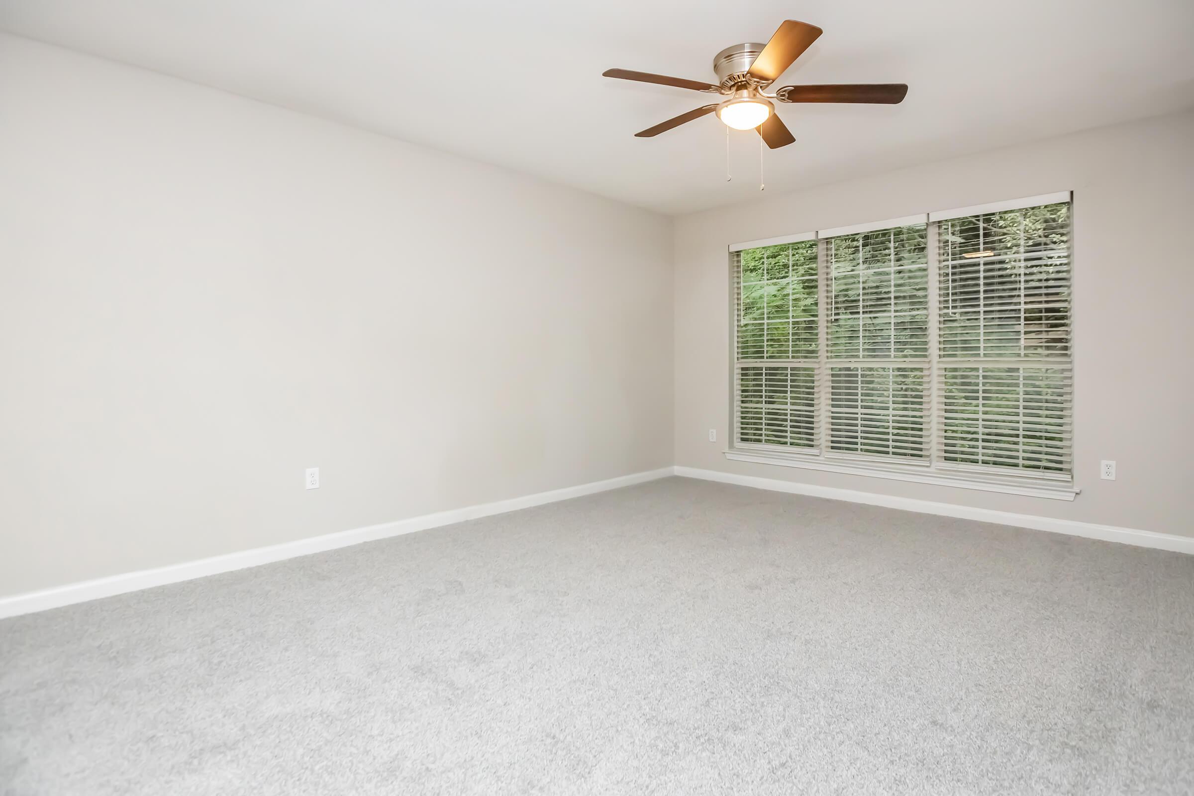 A spacious room with light gray carpet and beige walls. Features a ceiling fan with wooden blades and a large window with white blinds, allowing natural light to fill the space. The room is devoid of furniture, emphasizing its openness and potential for customization.