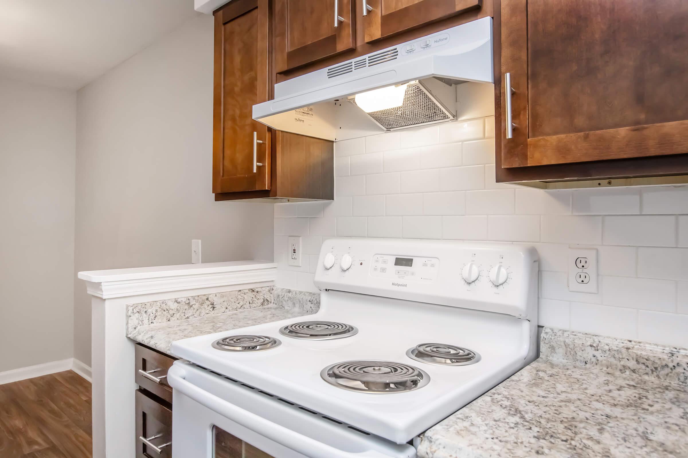 A close-up view of a modern kitchen featuring a white electric stove with four burners, a range hood above, and wooden cabinets. The countertops are made of granite, and the backsplash consists of white tiles, creating a clean and contemporary look. The kitchen has neutral-colored walls and a warm wooden floor.