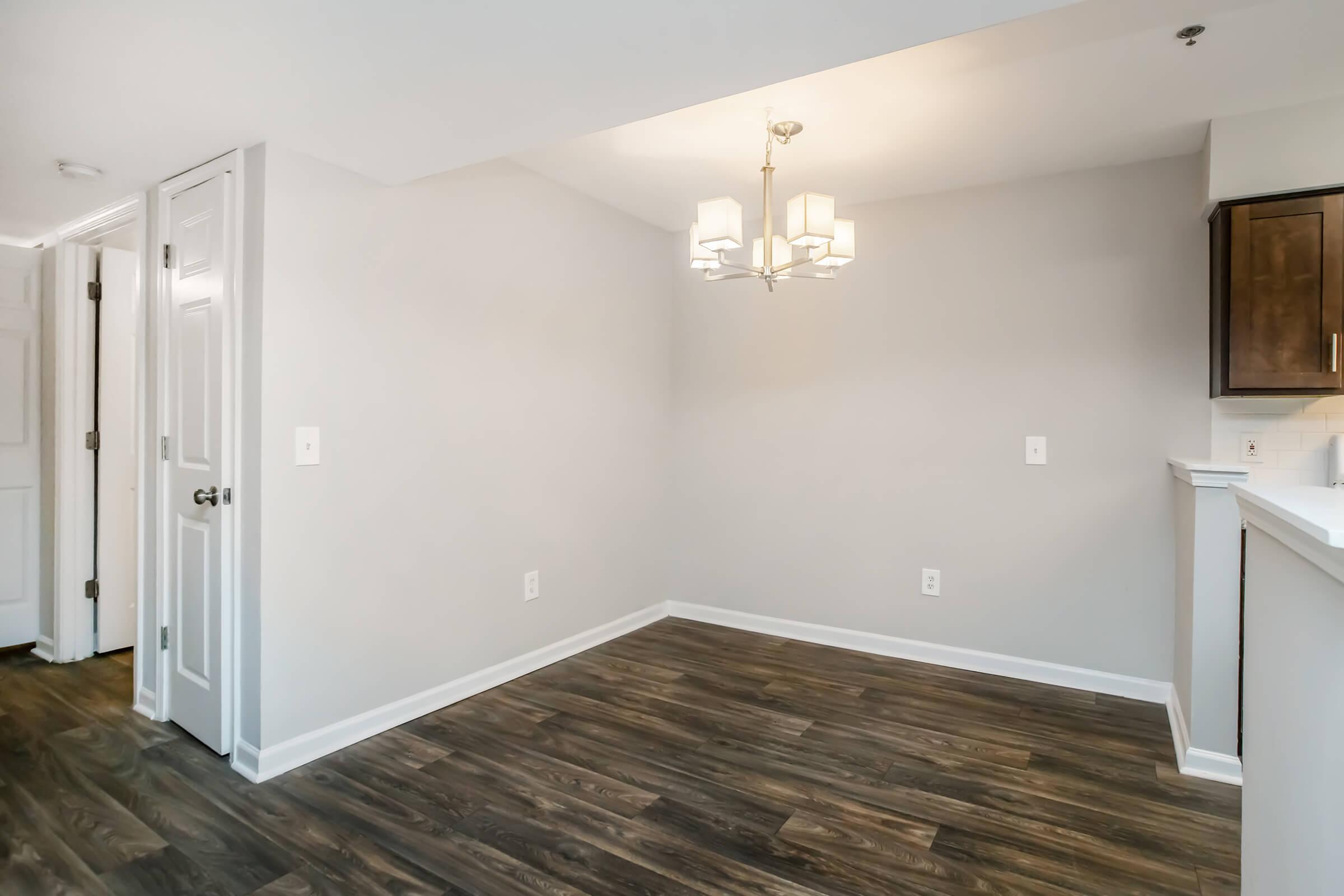 A modern, empty dining area featuring light gray walls and a stylish chandelier. The space has dark wooden flooring and an open layout connecting to a kitchen. There are two doors leading to other parts of the home, emphasizing the bright and spacious feel of the room.