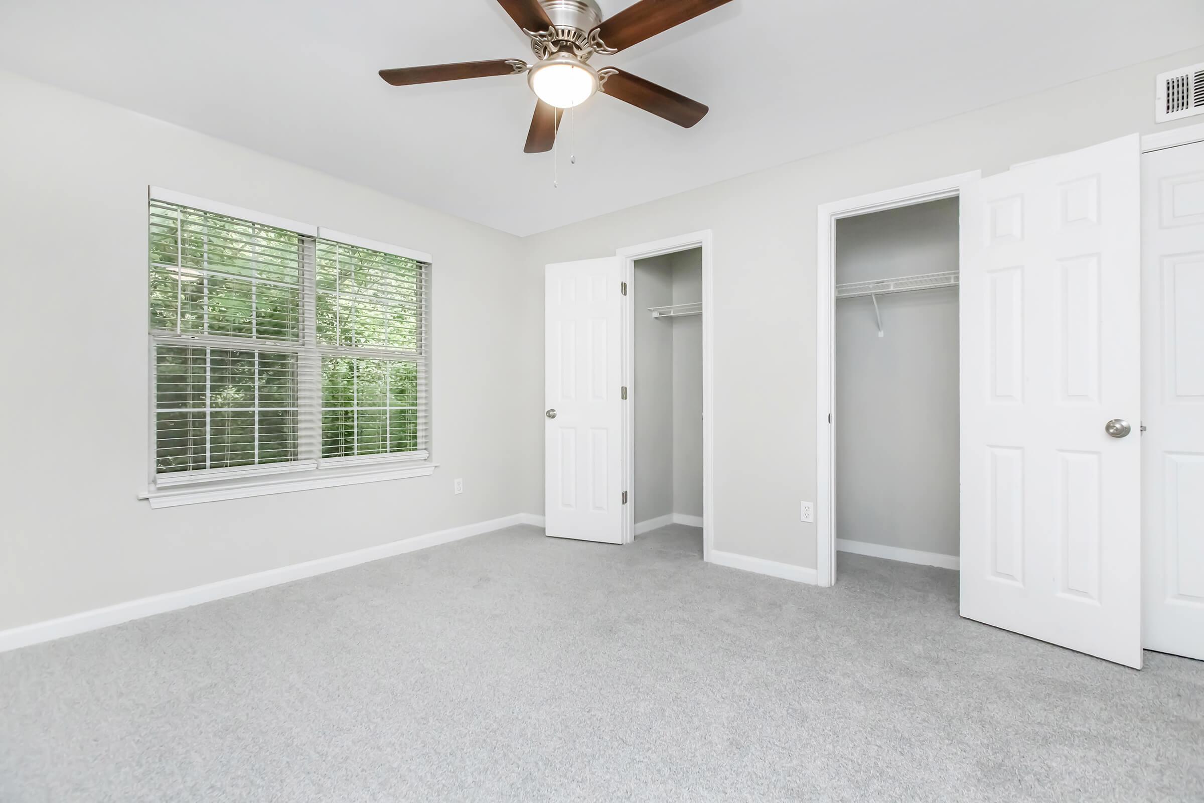 A spacious, light-filled bedroom featuring gray carpet, a ceiling fan, and a large window with blinds. There are two white double doors leading to a closet and another door to an empty closet space. The walls are painted a light color, creating a bright and inviting atmosphere.
