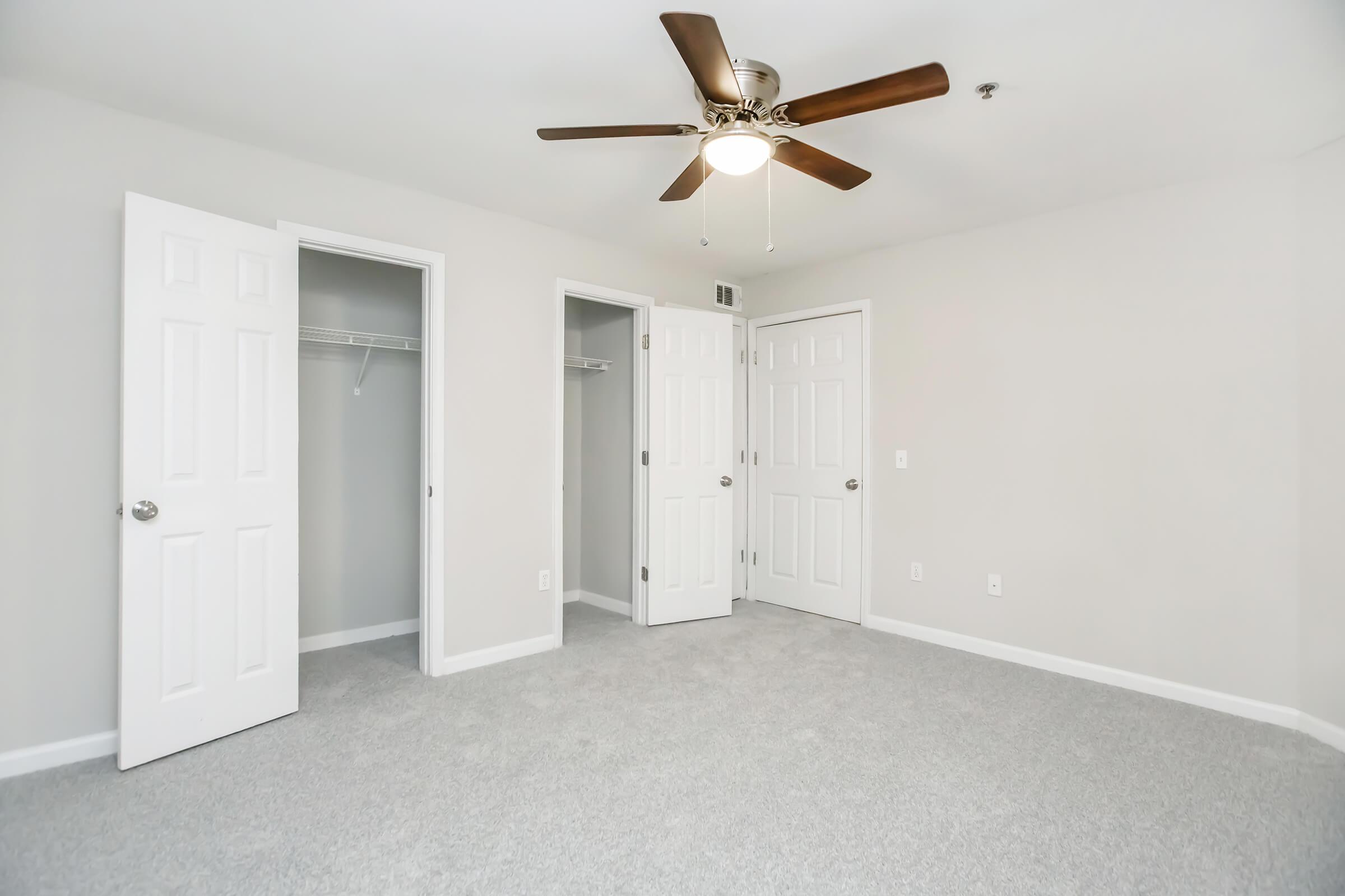 A spacious, light-colored room with gray carpet, featuring a ceiling fan. There are three white doors: one open revealing a closet, and two closed. The walls are painted a neutral hue, creating a clean and airy atmosphere.
