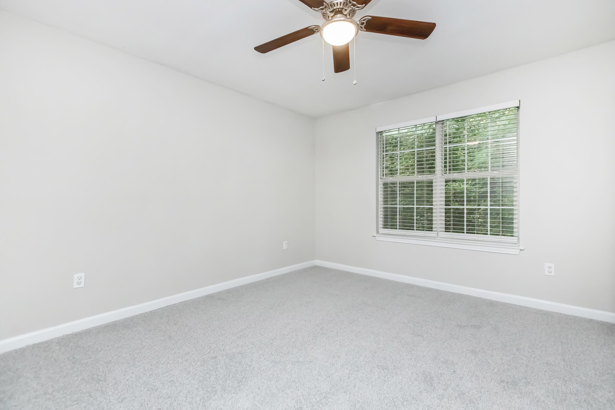 Empty room with light gray carpet, white walls, and a ceiling fan. A window with white blinds lets in natural light, showing greenery outside. The space is uncluttered and ready for furnishing, creating a neutral and inviting atmosphere.