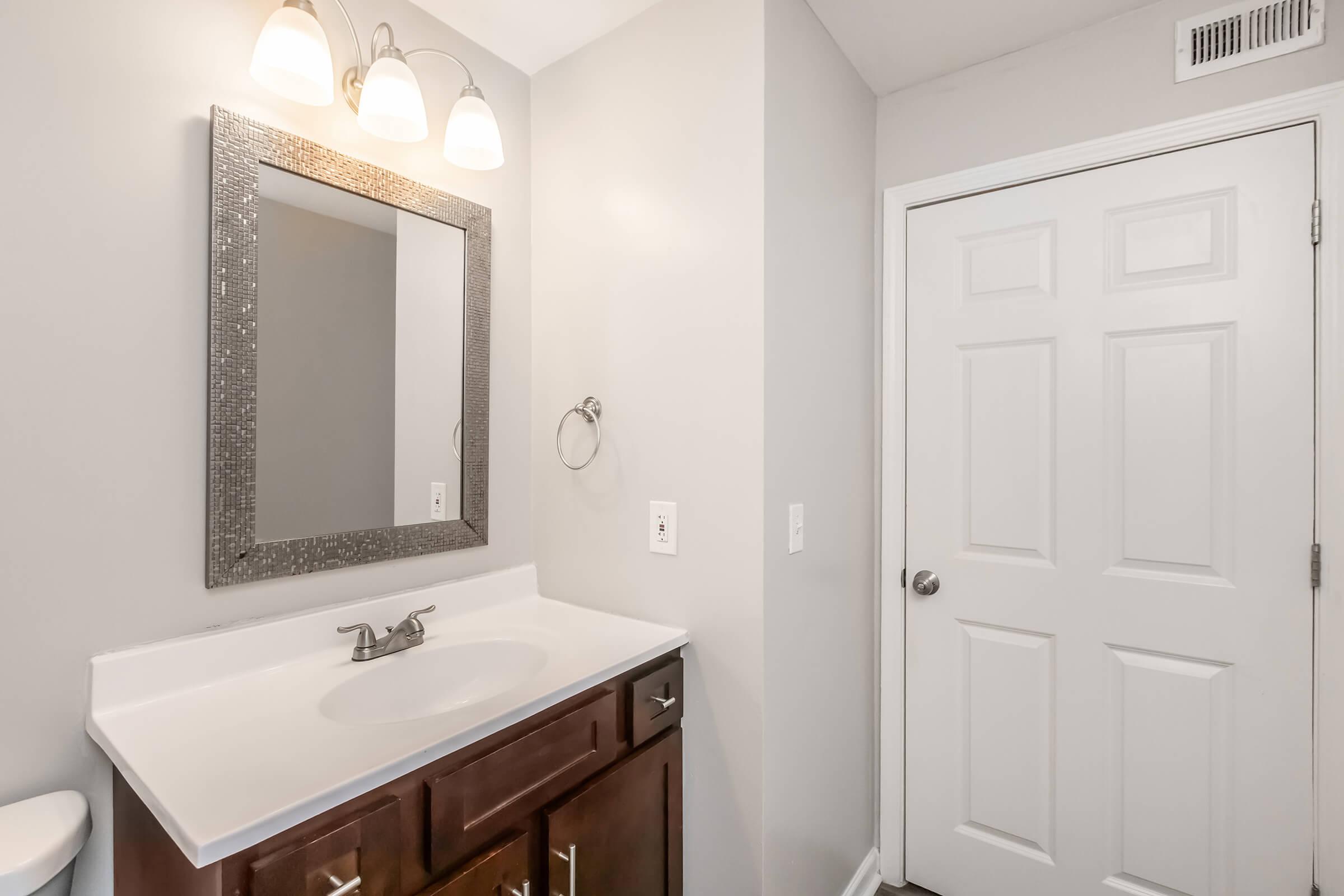 A clean and modern bathroom featuring a white countertop sink with a faucet, a rectangular mirror with a decorative frame, light fixtures above the mirror, and a gray wall. A white door is visible on the right, enhancing the minimalist aesthetic of the space.