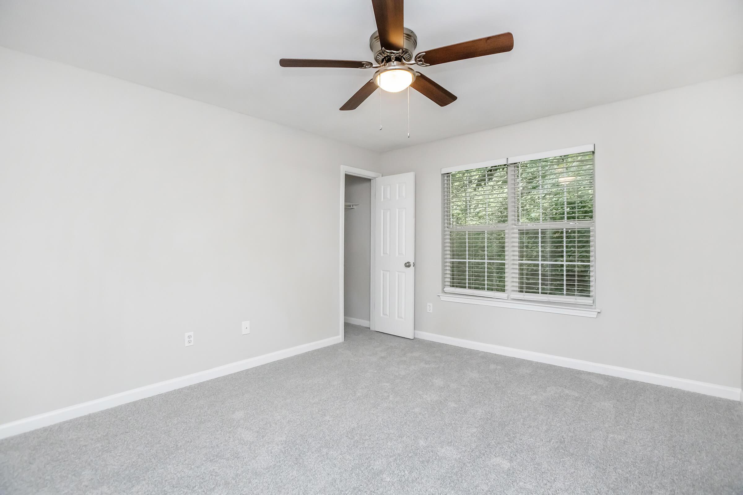 A spacious, empty bedroom with light gray carpet, a ceiling fan with wooden blades, and white walls. There's a window with multiple panes allowing natural light and a door leading to a closet. The room is well-lit and has a clean, neutral aesthetic.