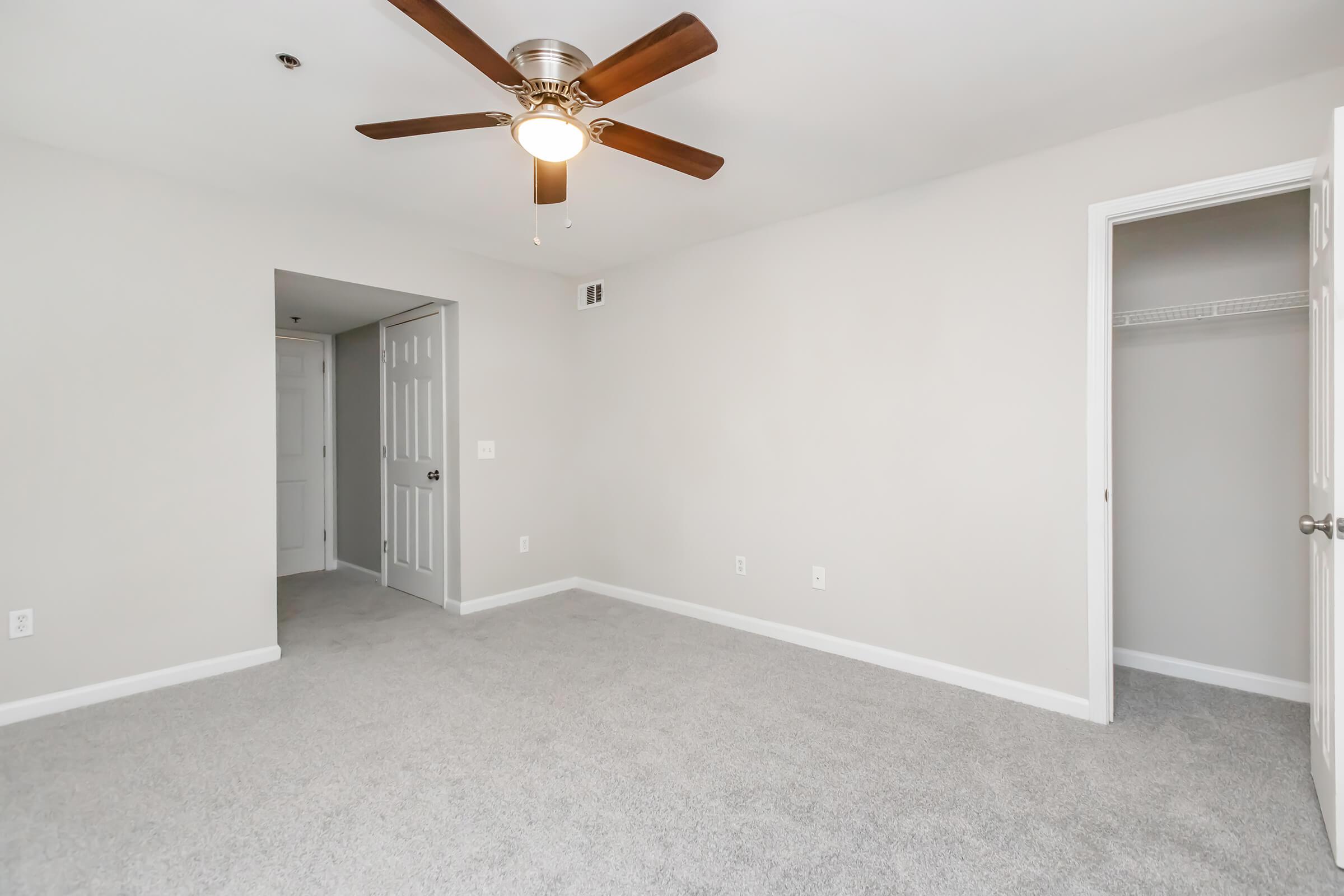 A spacious, empty bedroom featuring light gray walls and carpet. There is a ceiling fan with wooden blades and a light fixture. A doorway leads to another room, while a closet with sliding doors is visible. The overall atmosphere is bright and neutral, suitable for various decor styles.
