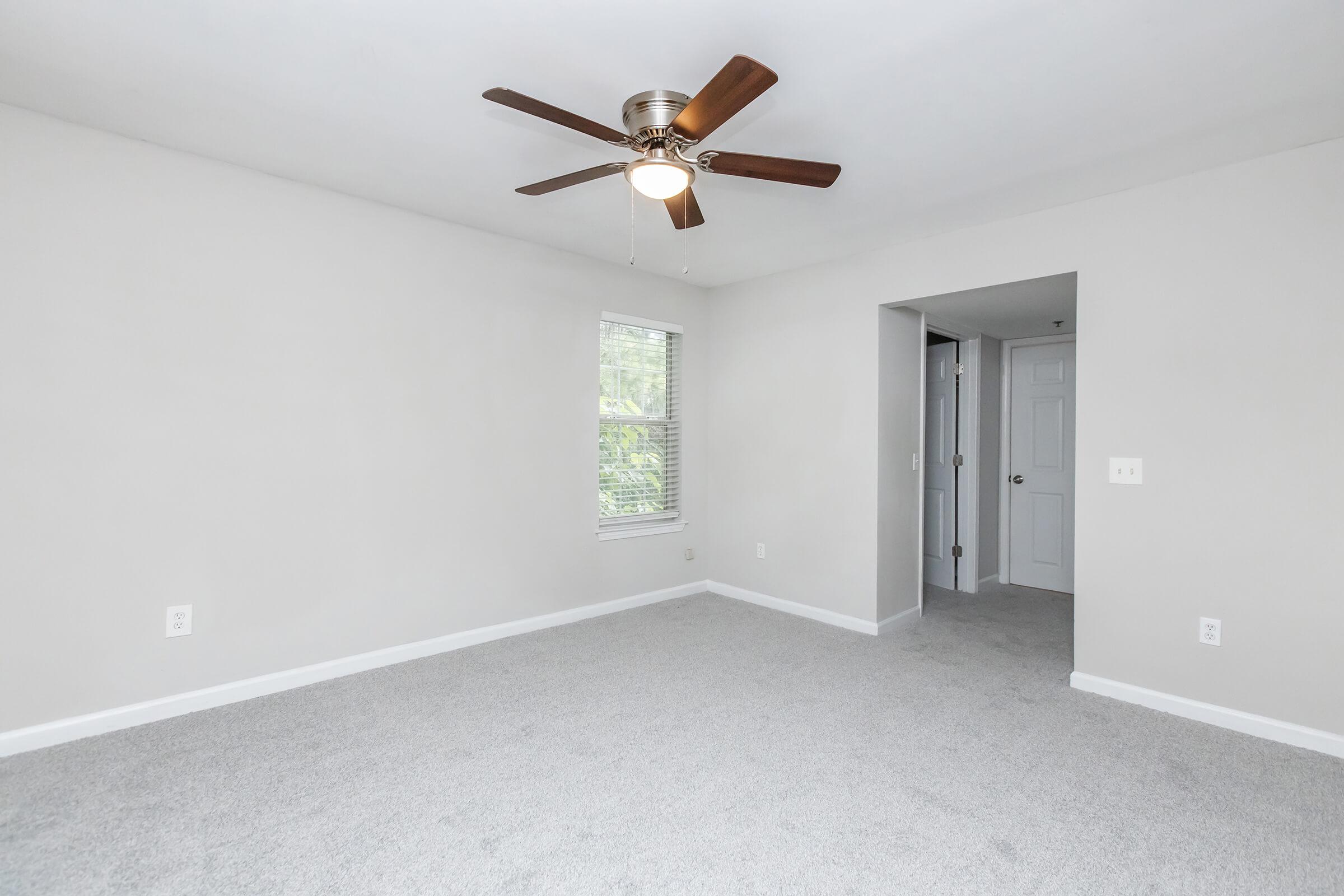 Empty room with light gray walls and carpet. A ceiling fan with wooden blades hangs in the center. There is a window on the left side, allowing natural light, and an entrance to another room on the right. The space is well-lit and minimalist, suitable for various furniture arrangements.