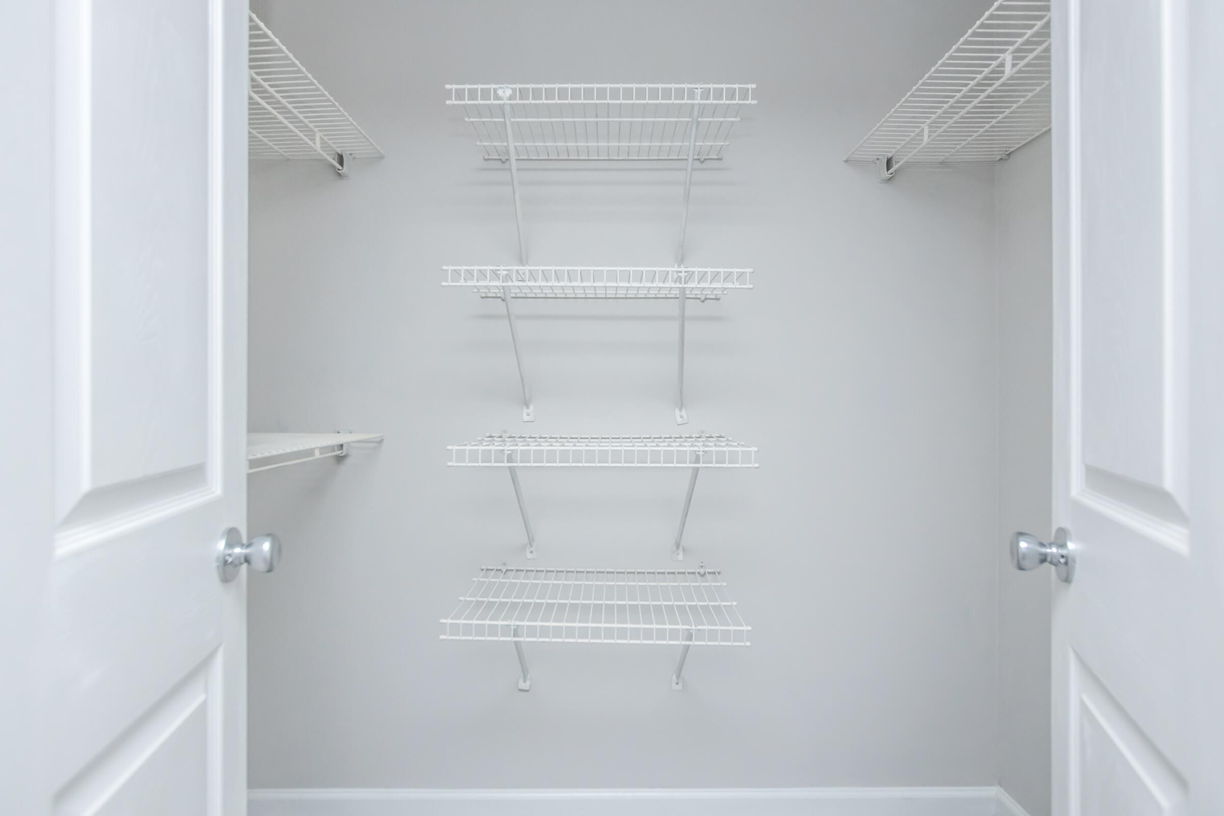 Empty closet with white wire shelving mounted on light gray walls. The closet has two white doors slightly ajar, revealing three horizontal shelves and one vertical shelf, all supported by metal brackets. The space appears clean and organized, awaiting storage items.