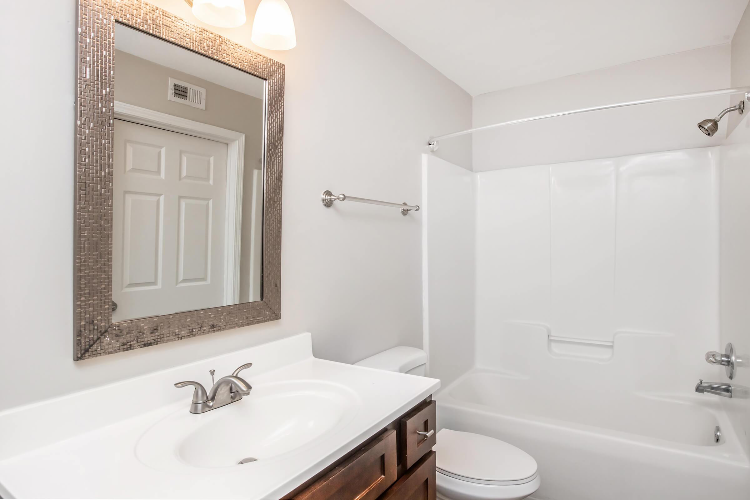 A clean and modern bathroom featuring a white bathtub and shower, a white vanity with a sink, and a large rectangular mirror above the sink. The walls are painted light gray, and there is a towel bar on the wall. A door is visible in the background, leading to another room.