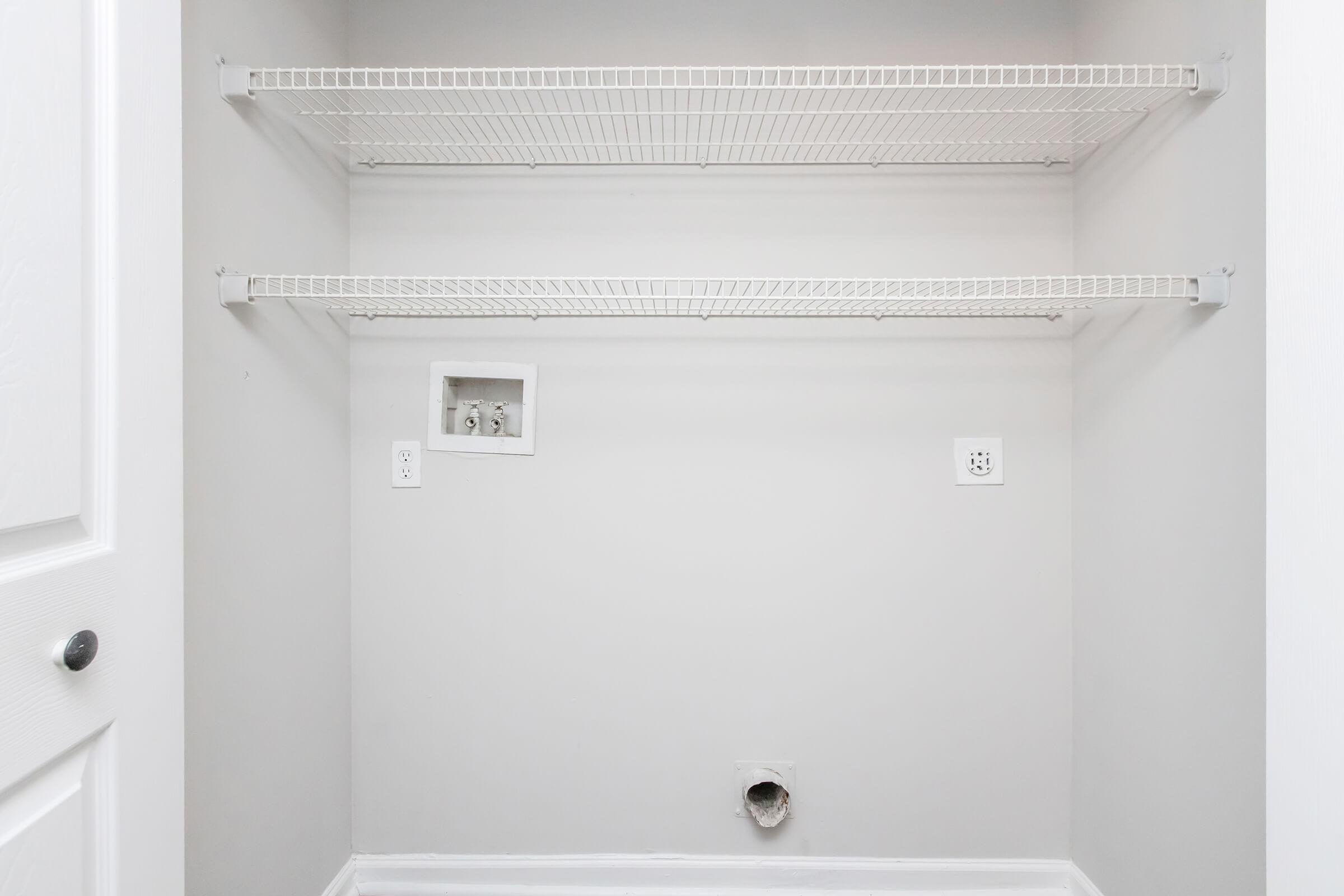 Empty closet with two wire shelves against a light gray wall. There are electrical outlets on either side, and a small cubby or opening in the wall at the back. The floor is bare, and there are no items or decorations inside the closet.