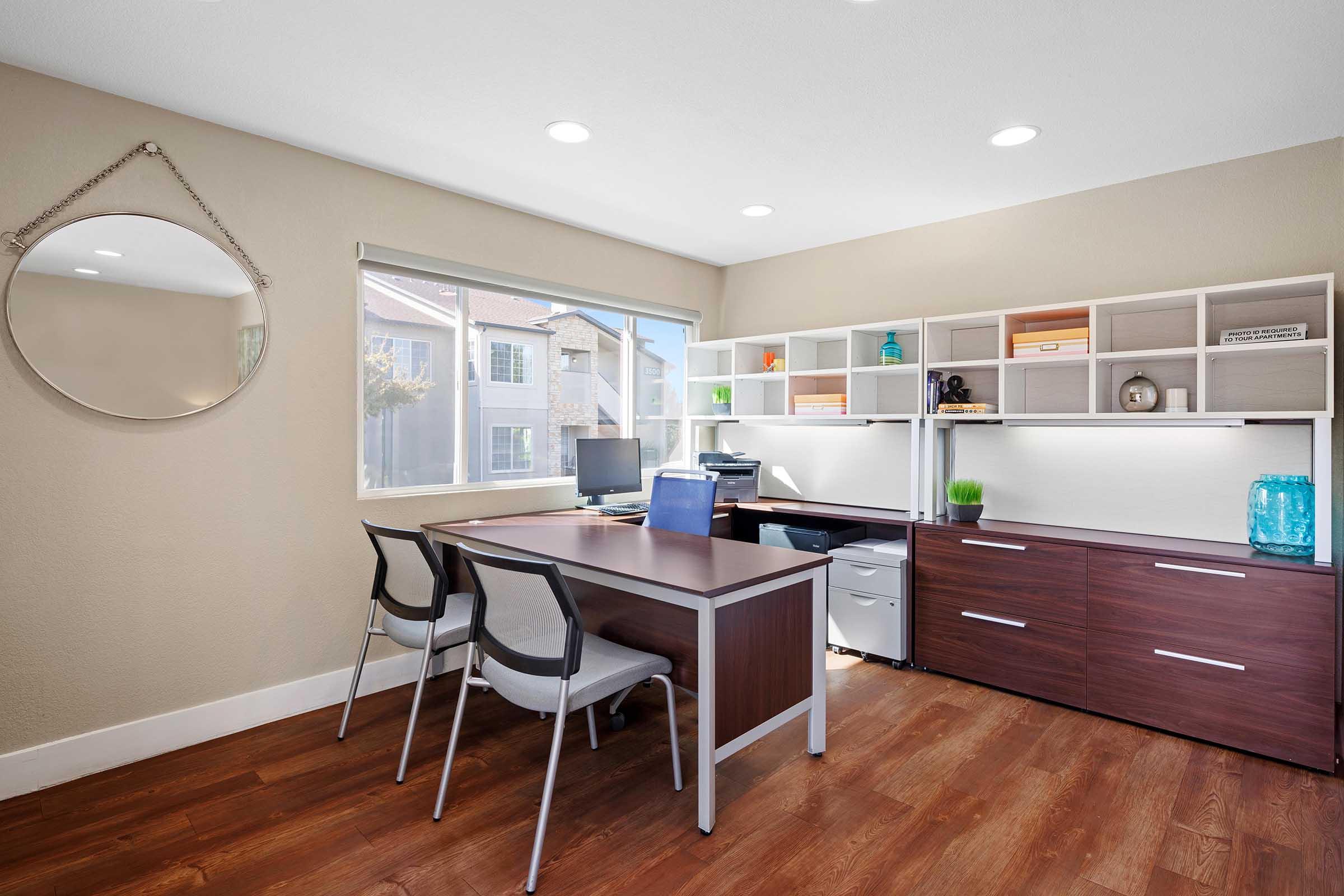 Modern office space featuring a wooden desk with two chairs, a computer setup, and a storage unit with shelves. A round mirror hangs on the wall, and there’s a window allowing natural light, showcasing a view of the outdoors. The room has a clean, minimalistic design with warm wooden flooring.