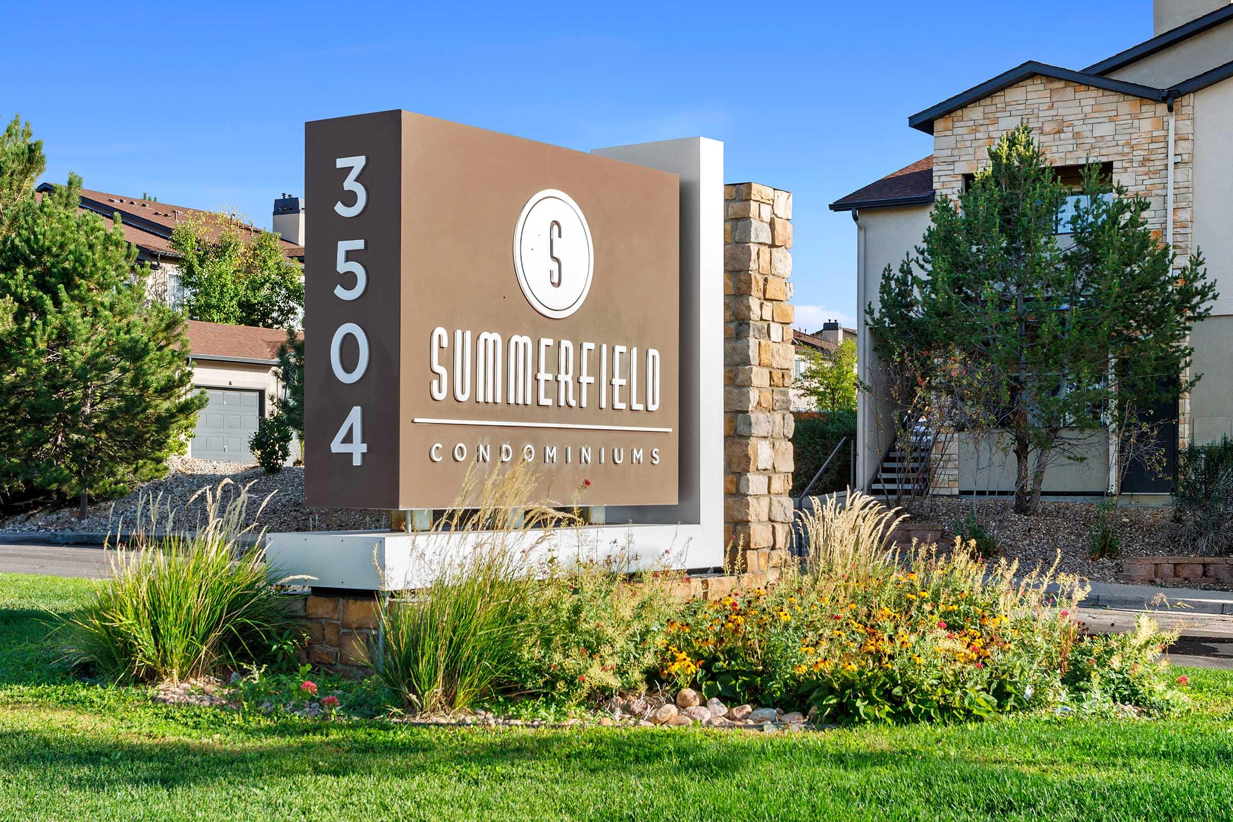 Signage for Summerfield Condominiums, displaying the address "3504." Surrounded by lush green grass and colorful flowers, with a clear blue sky above. The structure features a modern design with stone and wood elements.