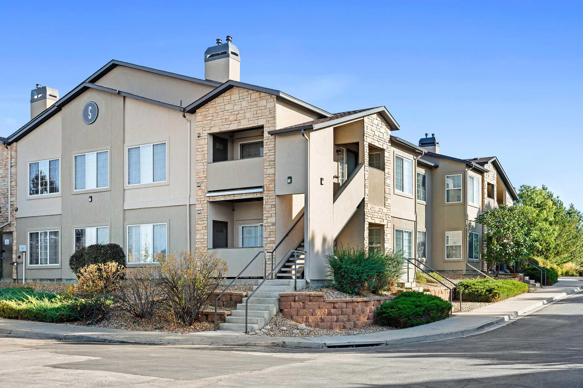 A two-story apartment building featuring a stone and stucco exterior. It has multiple windows, a set of stairs leading to the entrance, and landscaped shrubs and trees surrounding it. The sky is clear and blue, creating a bright and inviting atmosphere.