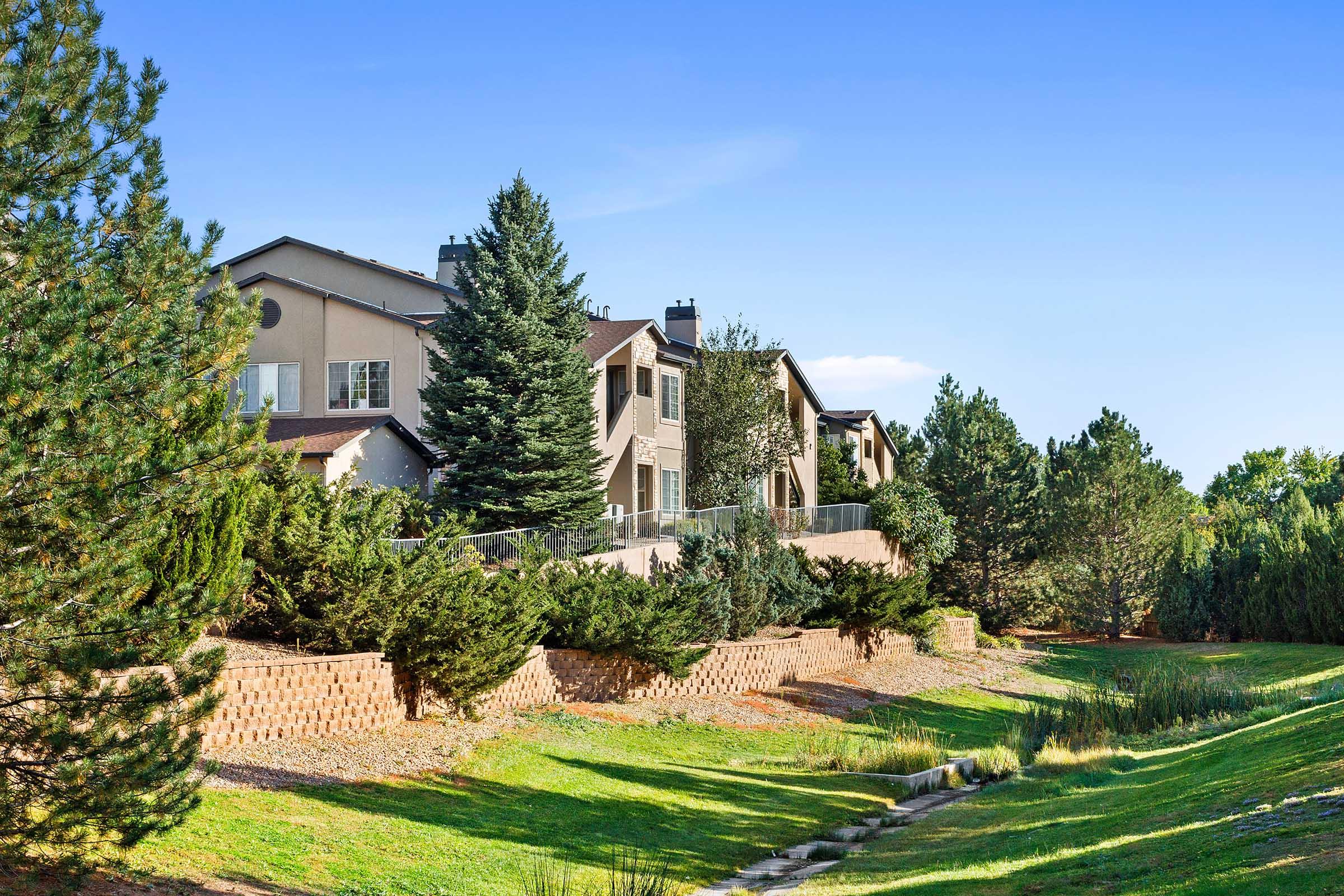 Row of modern houses surrounded by lush greenery and tall trees, with a clear blue sky in the background. The landscaping features a well-maintained lawn and a gentle slope leading to a water feature or stream. The scene conveys a peaceful residential area.