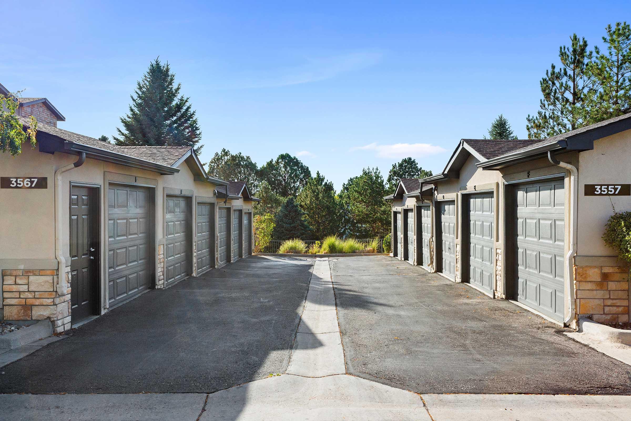 A row of garage doors with address numbers 3567 and 3557, set against a clear blue sky and surrounded by trees. The paved driveway leads to the garages, creating a neat and organized appearance.