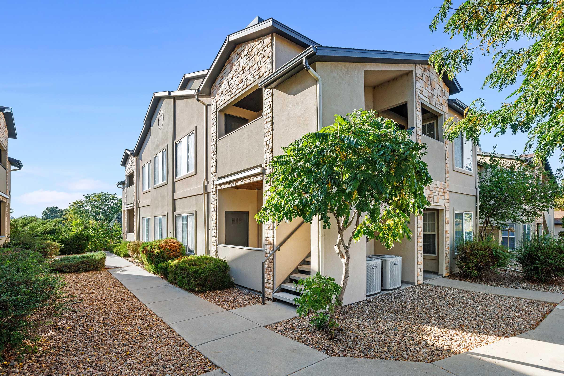 A multi-unit residential building with a modern design features multiple balconies and windows. The structure is surrounded by well-maintained landscaping, including trees and shrubs. A pathway leads to the entrance, with decorative stone accents on the exterior.