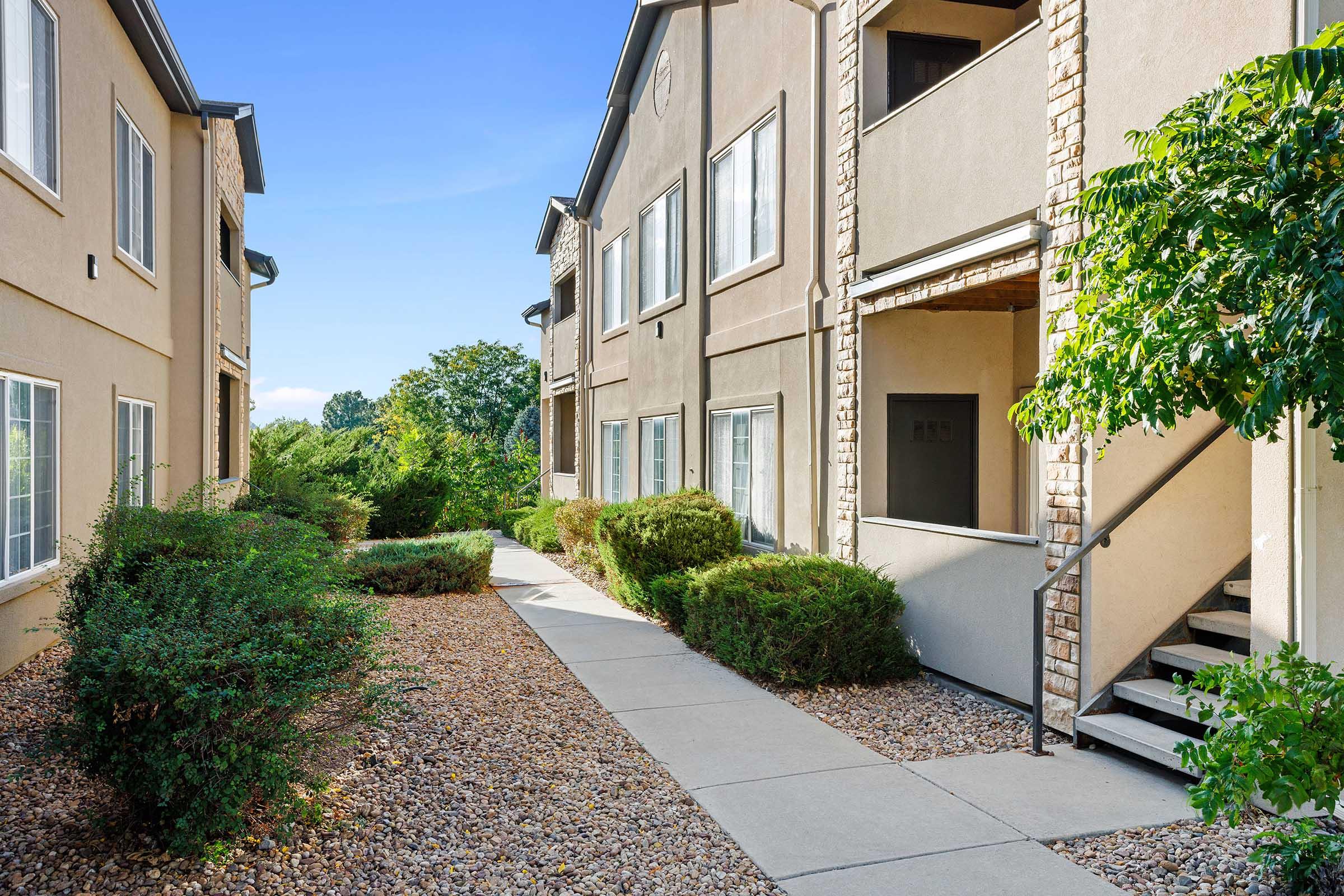 View of an apartment complex featuring a landscaped path lined with shrubs, leading to two-story beige buildings with balconies and stone accents. The clear blue sky adds to the outdoor atmosphere, while the greenery complements the modern architecture.