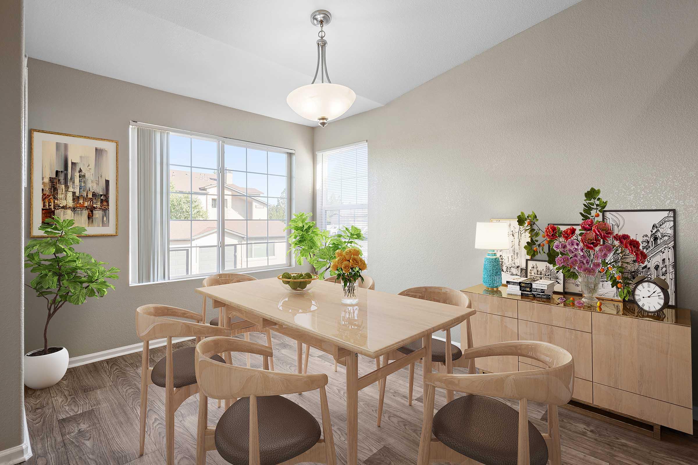 A modern dining room featuring a light wooden table surrounded by beige chairs. The room has a large window with natural light, green plants, and a floral arrangement on the table. A sideboard displays decorative items, including a clock and artwork, against a softly colored wall. A pendant light hangs from the ceiling.