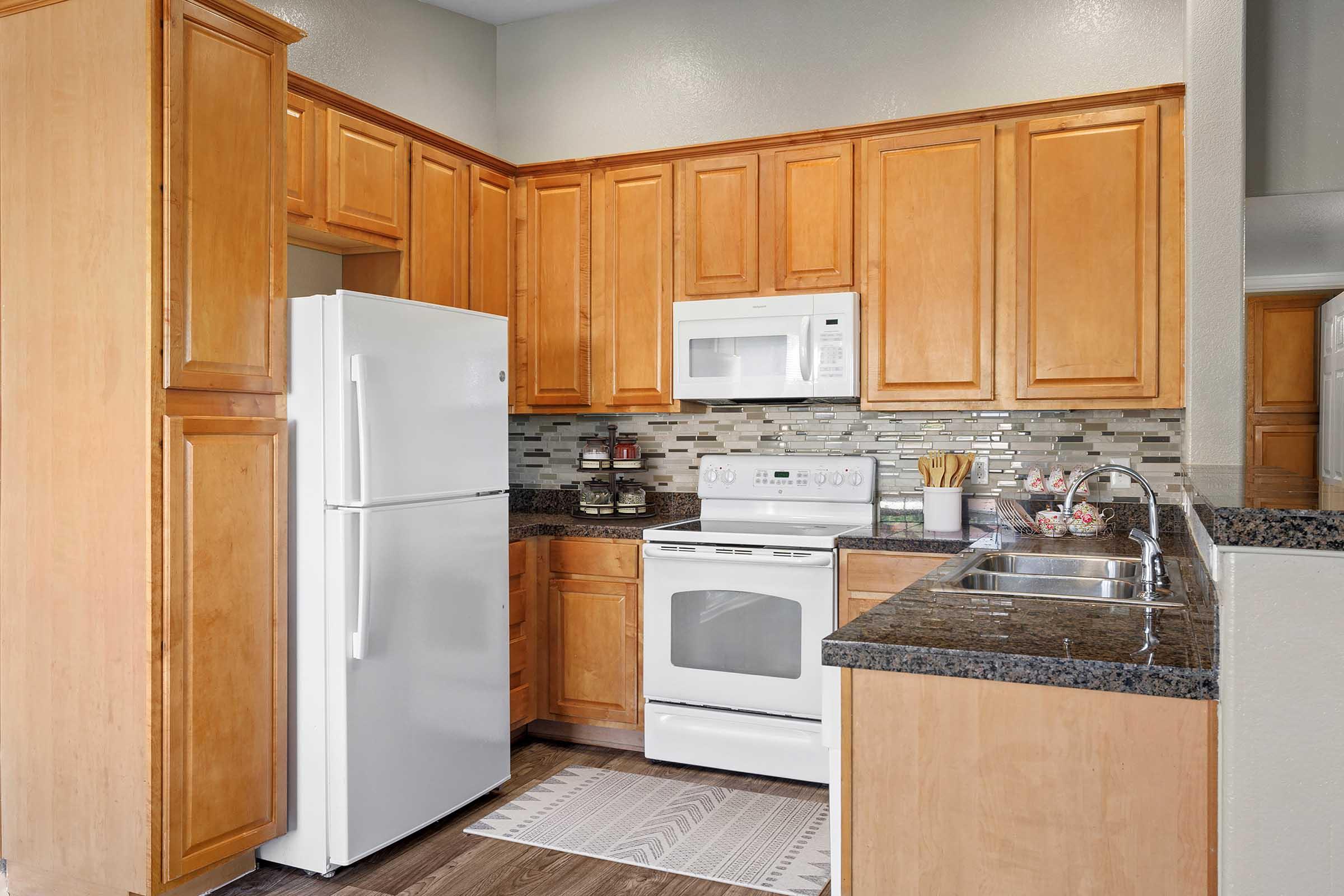 A modern kitchen featuring wooden cabinetry, a white refrigerator, a white stove, and a built-in microwave. The countertops are dark granite, and there's a sink next to the stove. The backsplash consists of glass tiles, and a small mat is placed on the floor.